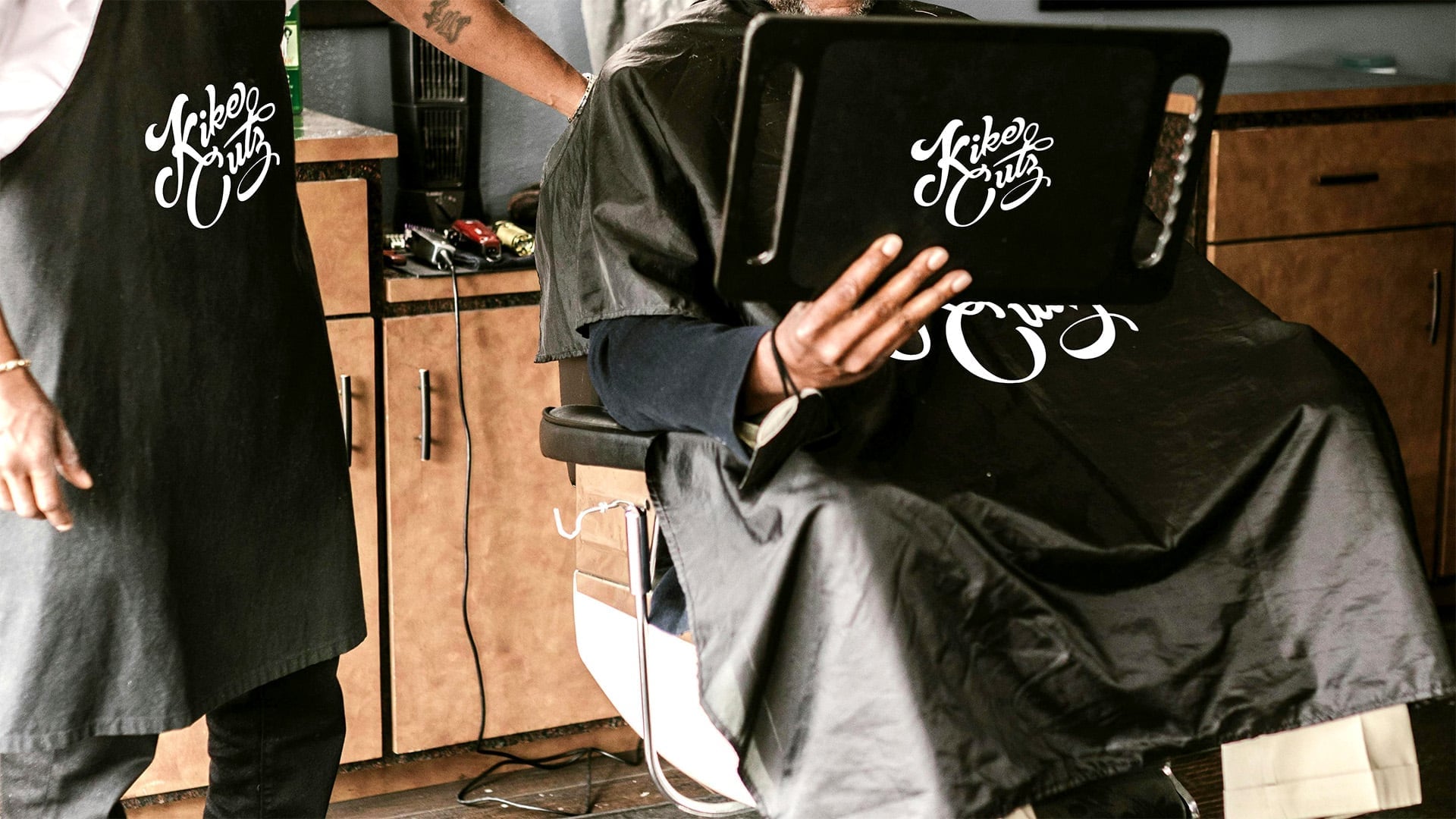 Barbershop branding mockup showing black apron and cutting cape with white Kike Cutz script logo, worn by barber and client in salon setting with wood cabinets visible