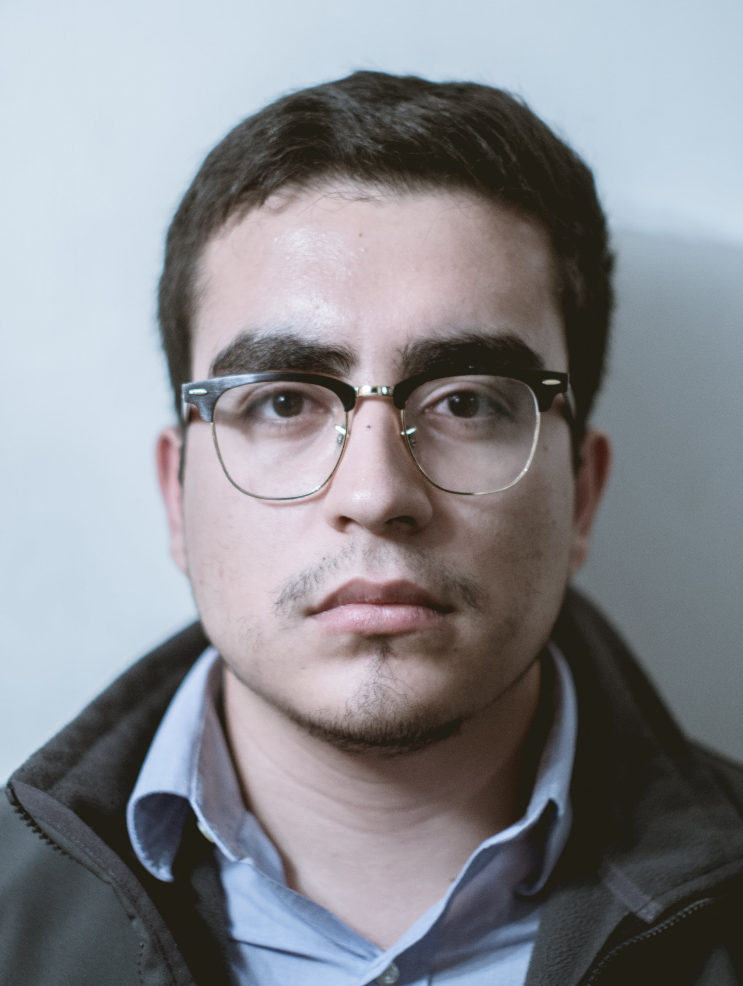 Formal portrait of a young man with glasses wearing a button-down shirt and jacket, neutral expression against a plain light background