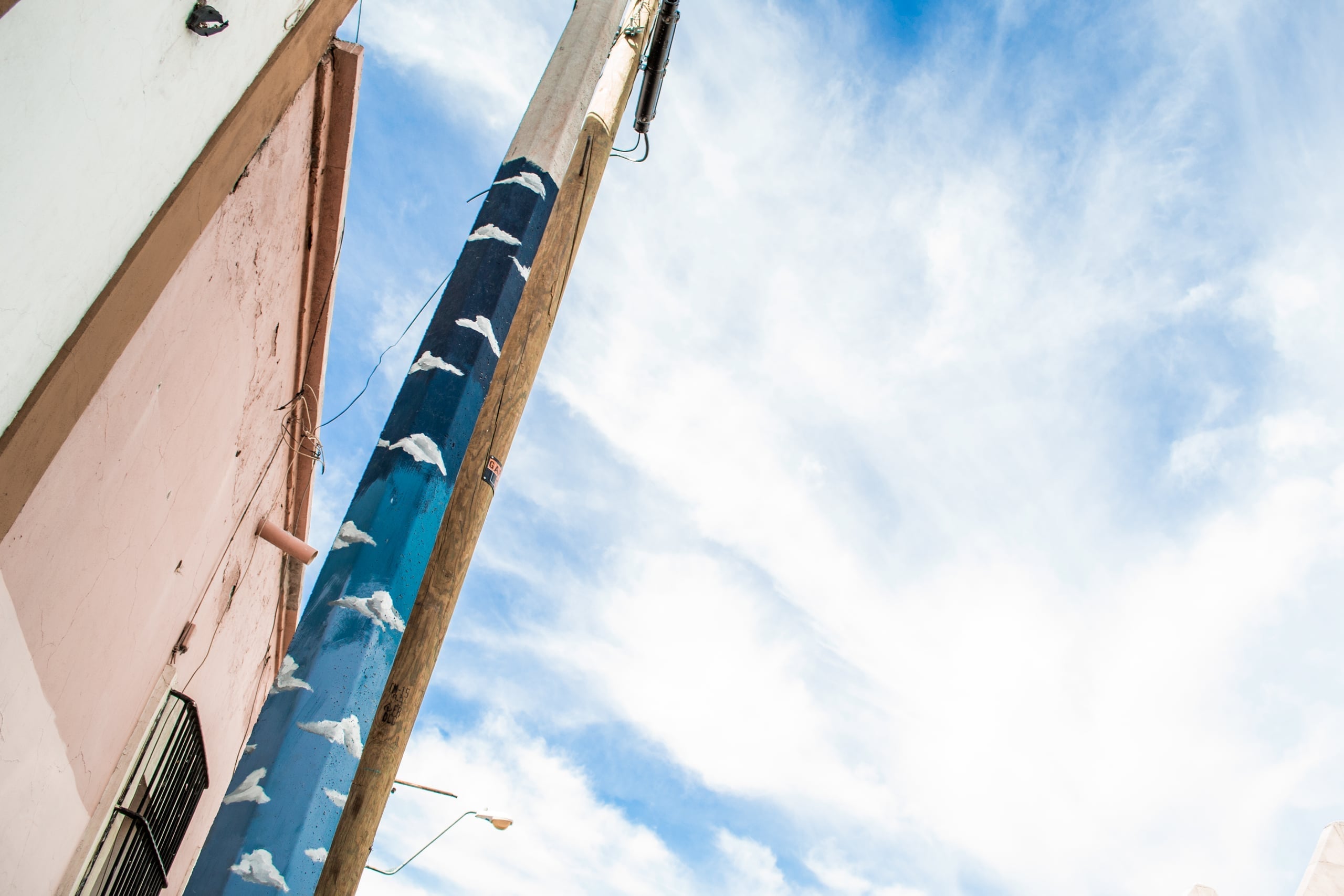 Looking up at colorful building facades - pink wall and blue pillar with white cloud murals painted on it - against a bright blue sky with wispy clouds