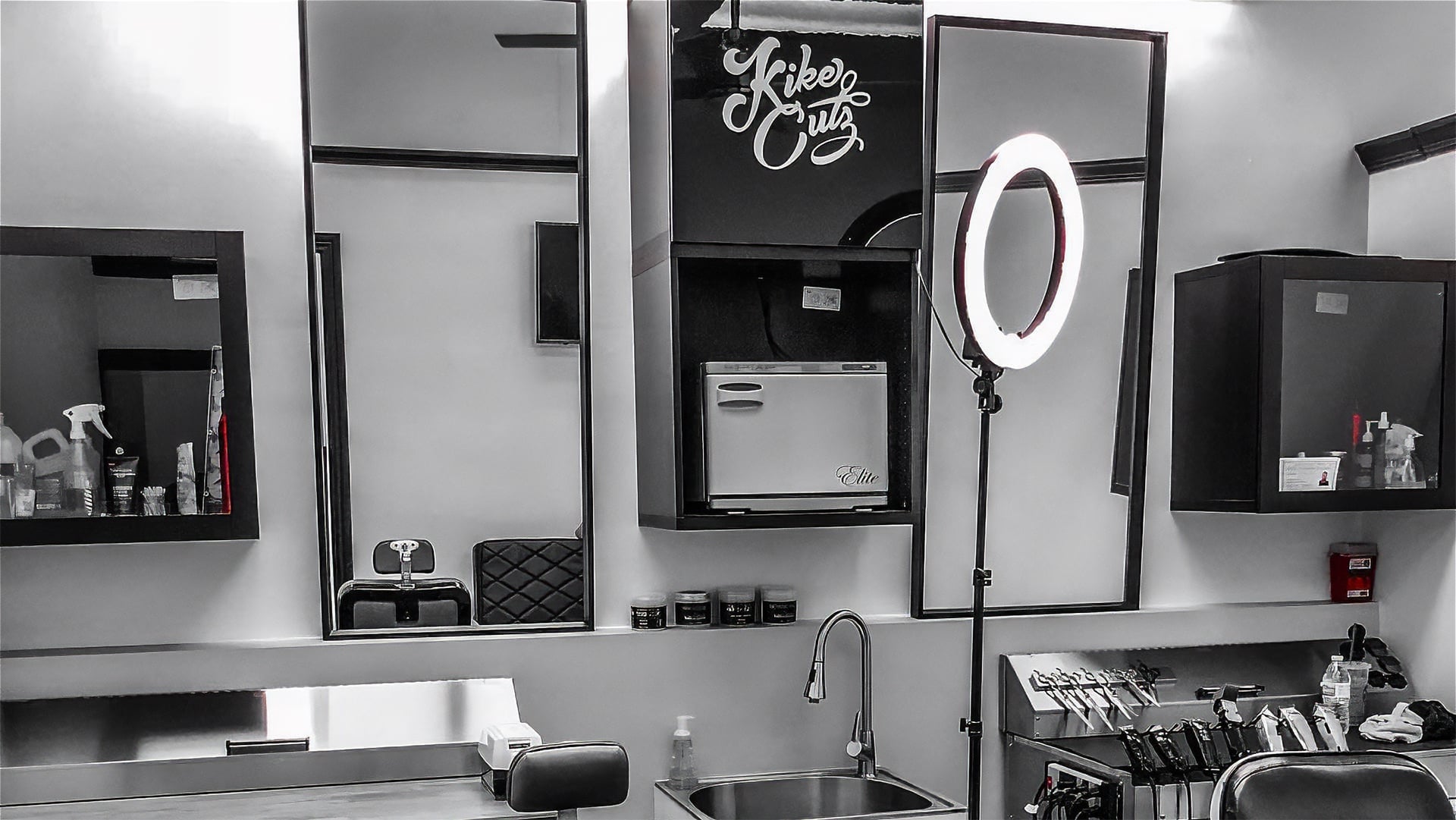 Modern barbershop interior in black and white showing Kike Cutz logo on wall, professional ring light, mirrors, cutting station with clippers, and towel warmer