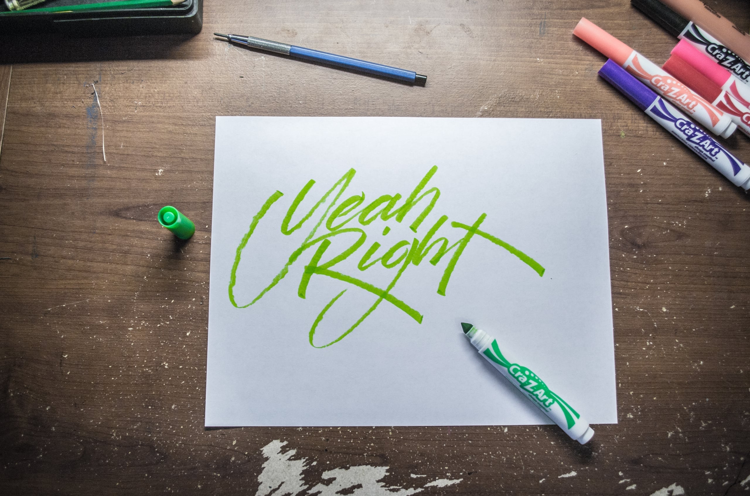 Flat lay photograph of green brush marker calligraphy reading 'Yeah Right' on white paper, surrounded by colorful markers on a wooden desk
