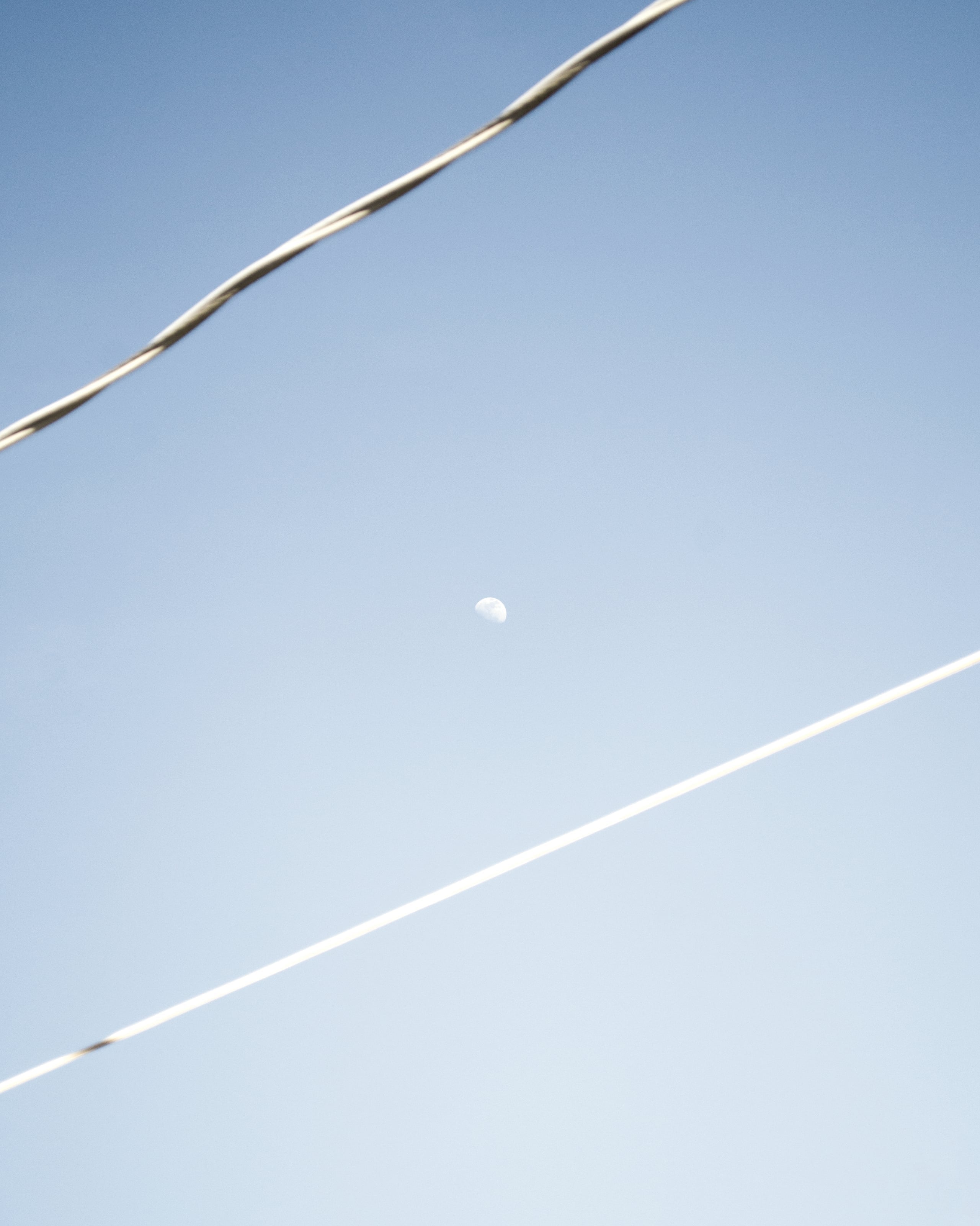 Minimalist photograph of a faint daytime moon centered between two diagonal power lines against a pale blue gradient sky