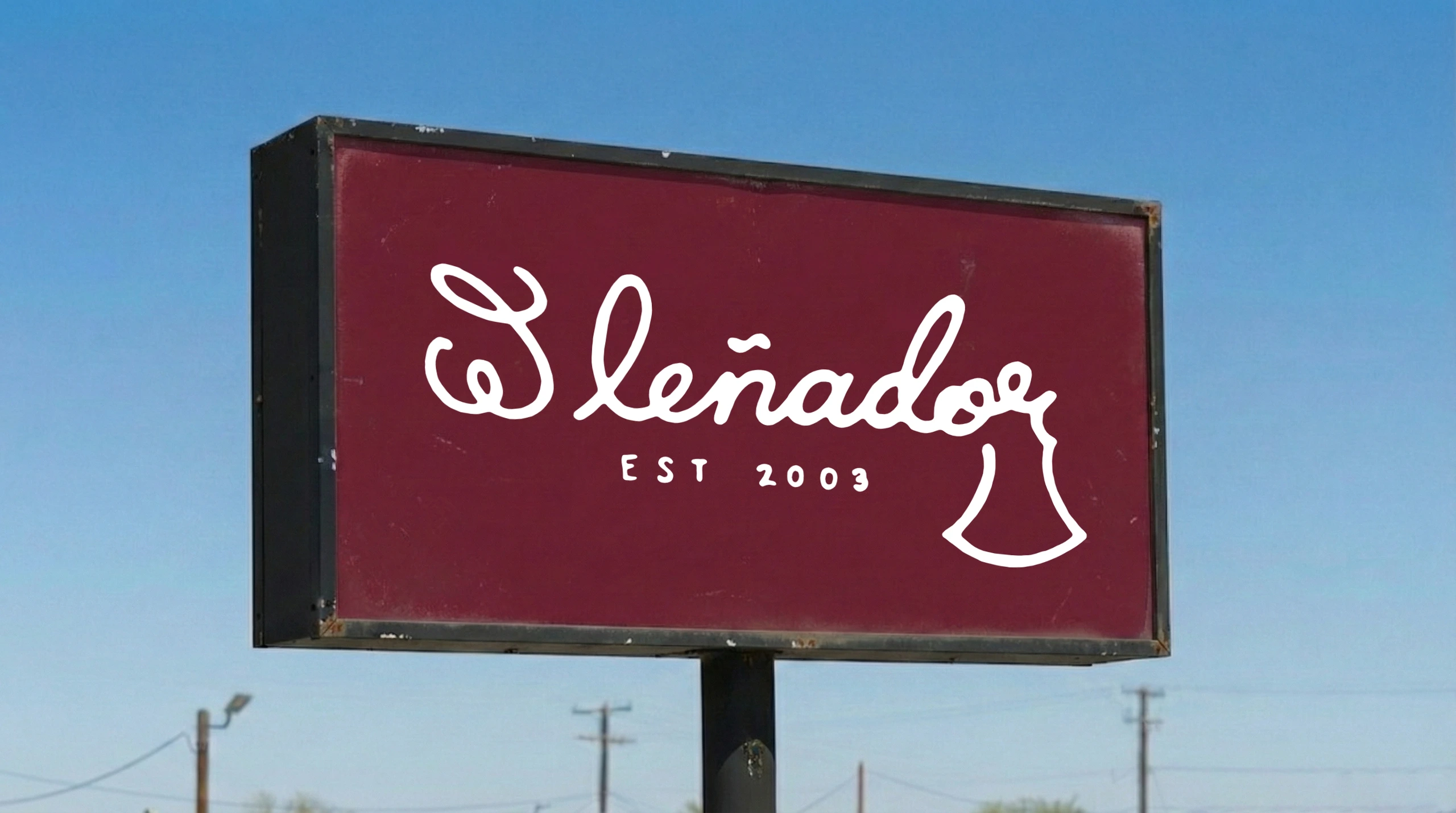 Outdoor rectangular sign with deep maroon background and black metal frame displaying El Lenador hand-lettered cursive logo in white with EST 2003 tagline against clear blue sky with power lines below