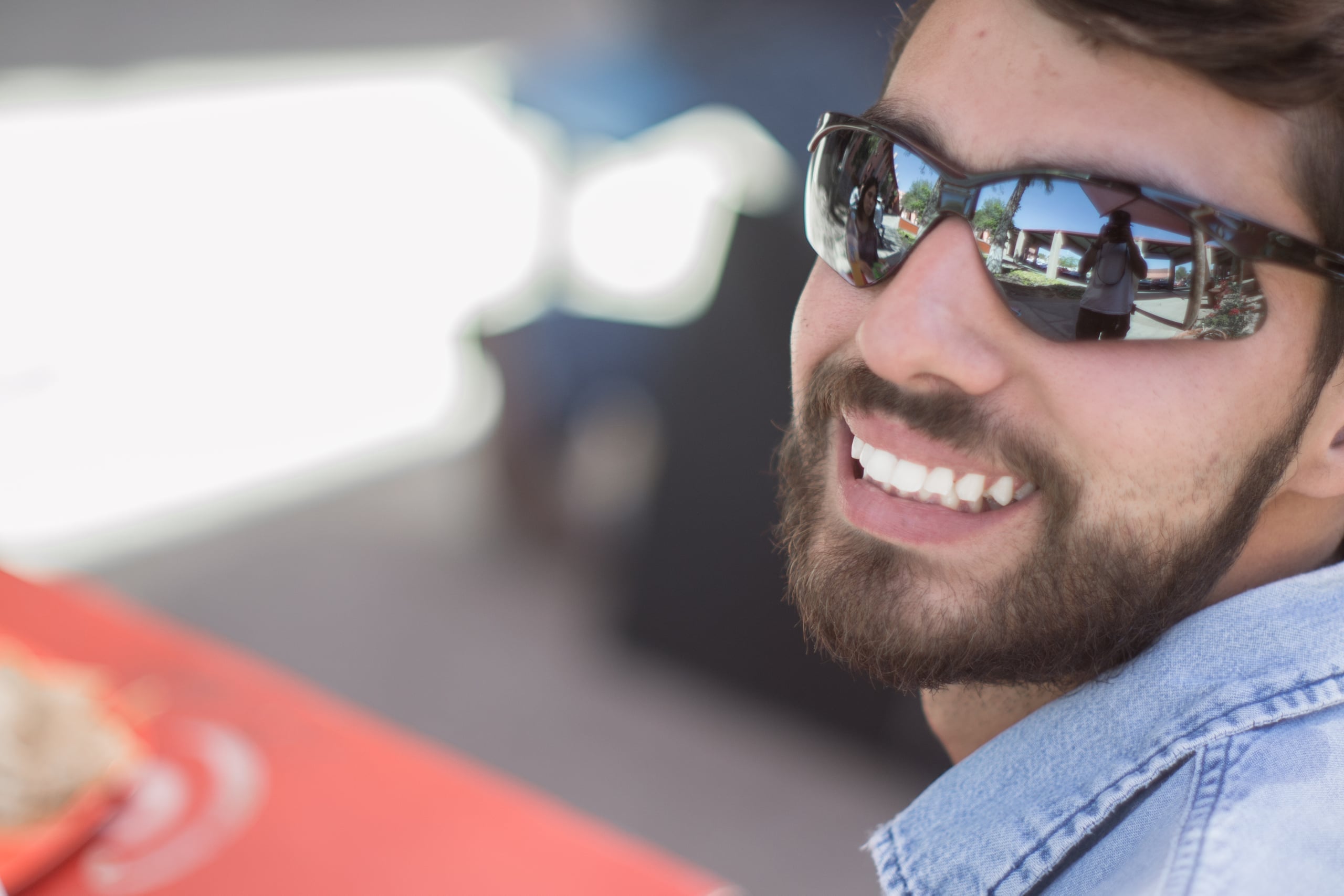 Close-up portrait of a smiling bearded man wearing reflective sunglasses and a denim jacket, with outdoor scenery reflected in the lenses