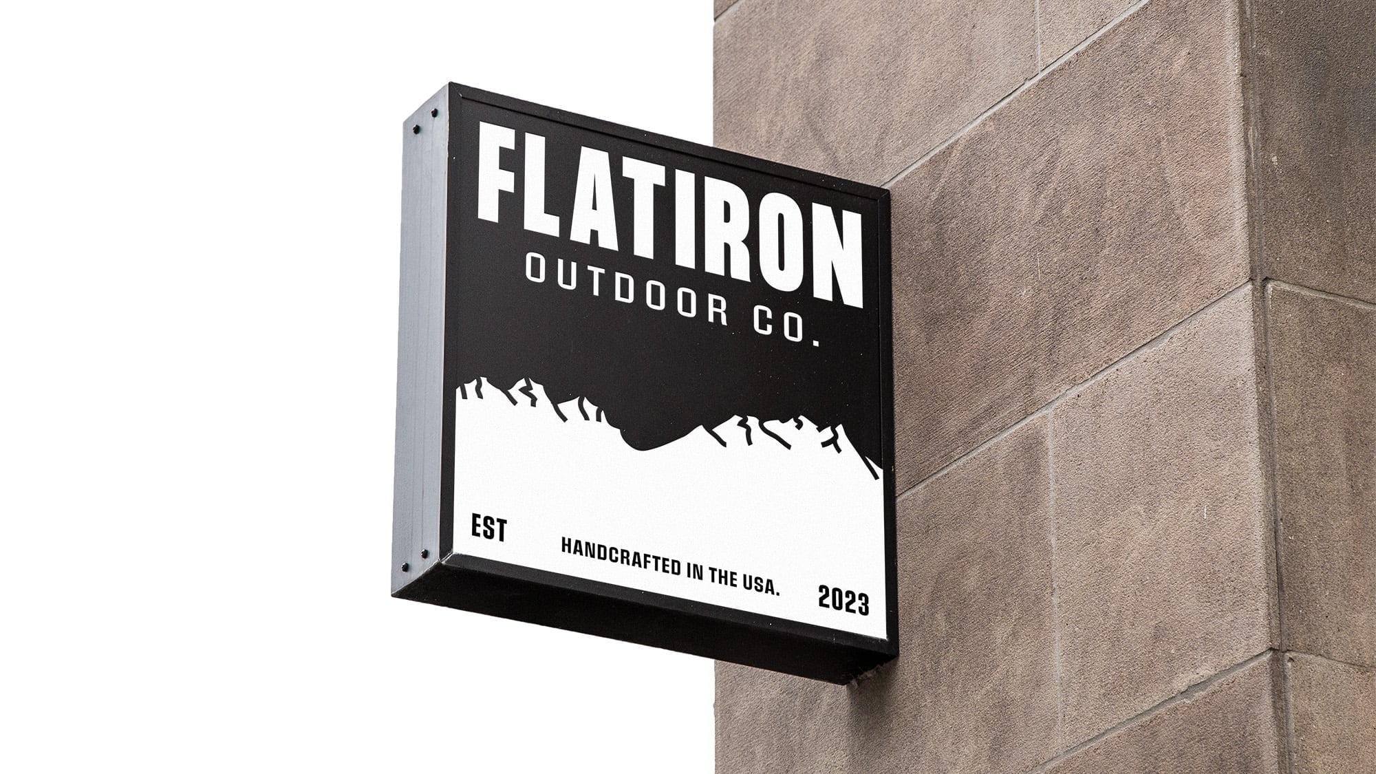 Square blade sign mounted on tan stone building wall displaying Flatiron Outdoor Co. logo with white mountain silhouette on black background and Handcrafted in the USA tagline.