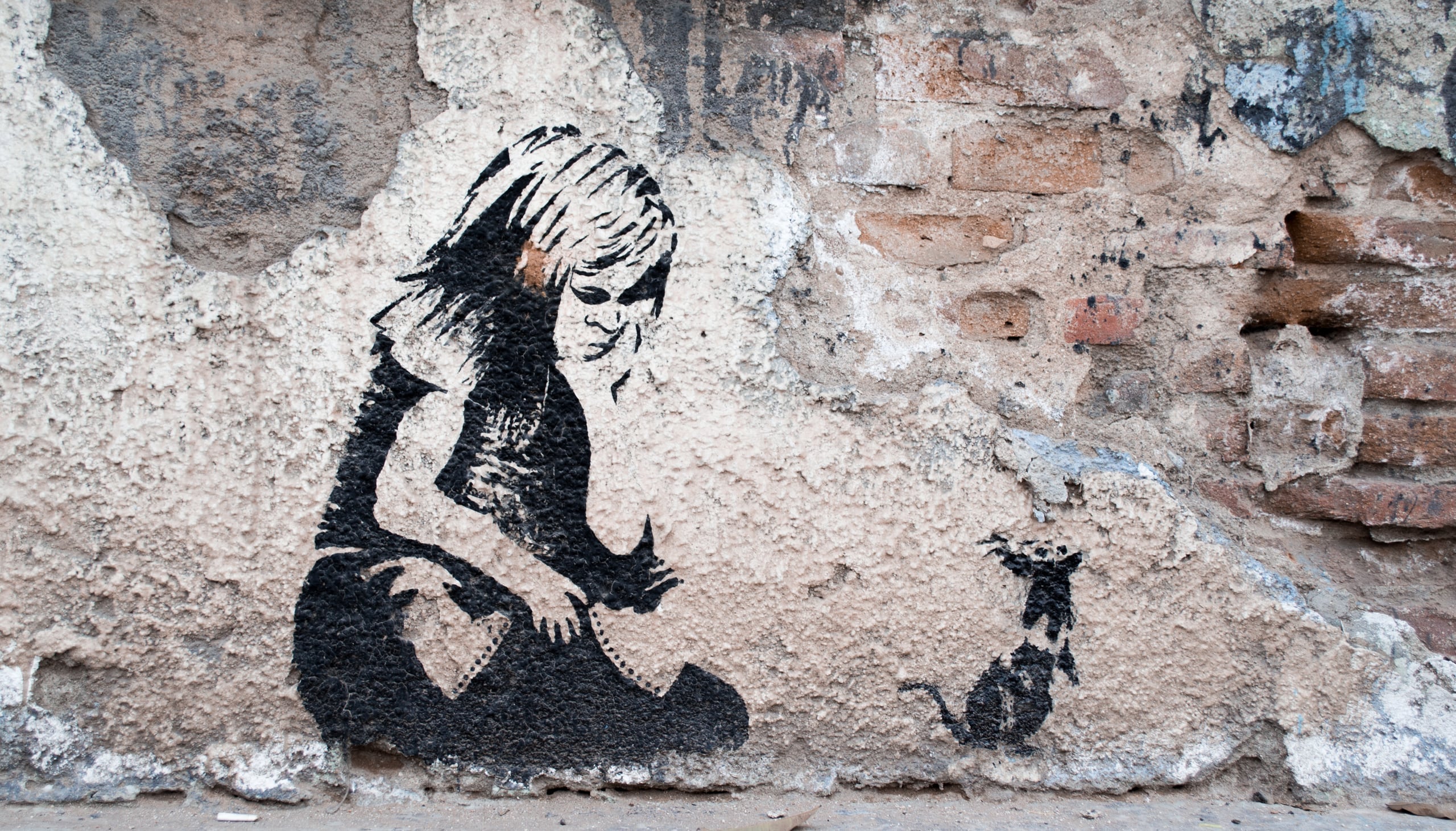 Stencil street art on a weathered brick and plaster wall depicting a seated girl reaching toward a small rat, in black spray paint