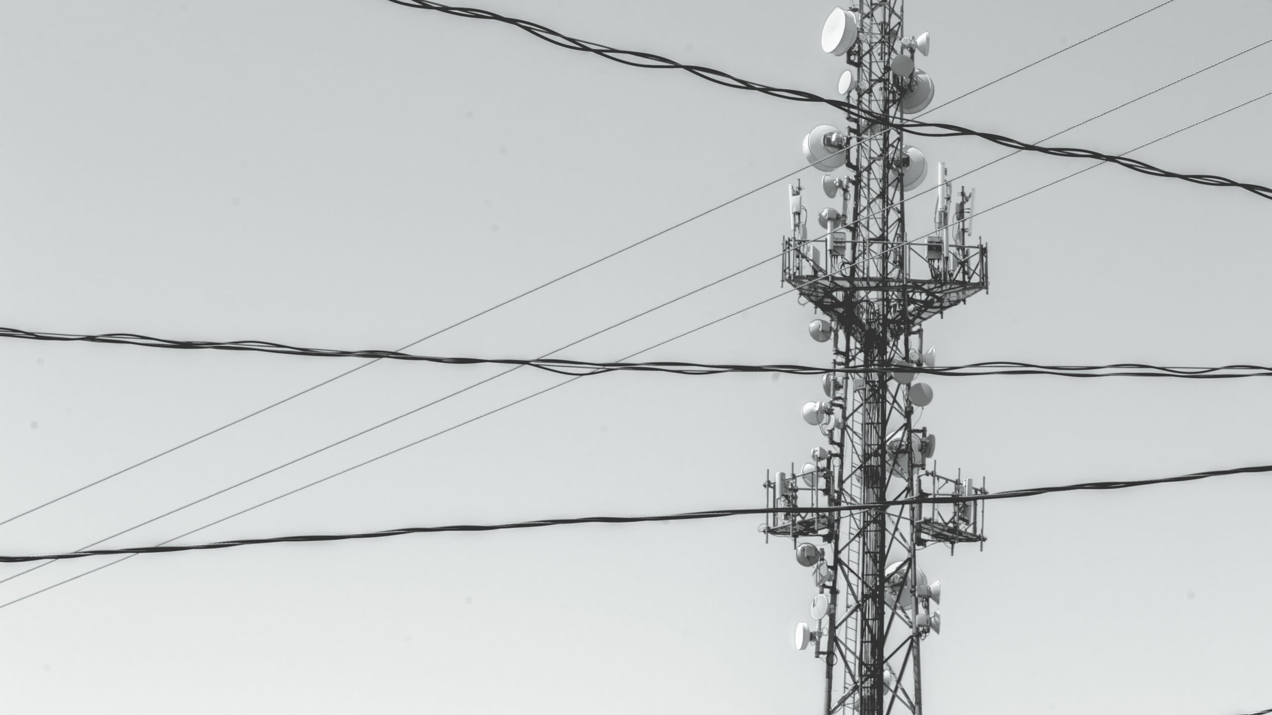 Telecommunications tower with multiple satellite dishes and antennas silhouetted against an overcast gray sky, with power lines crossing the frame