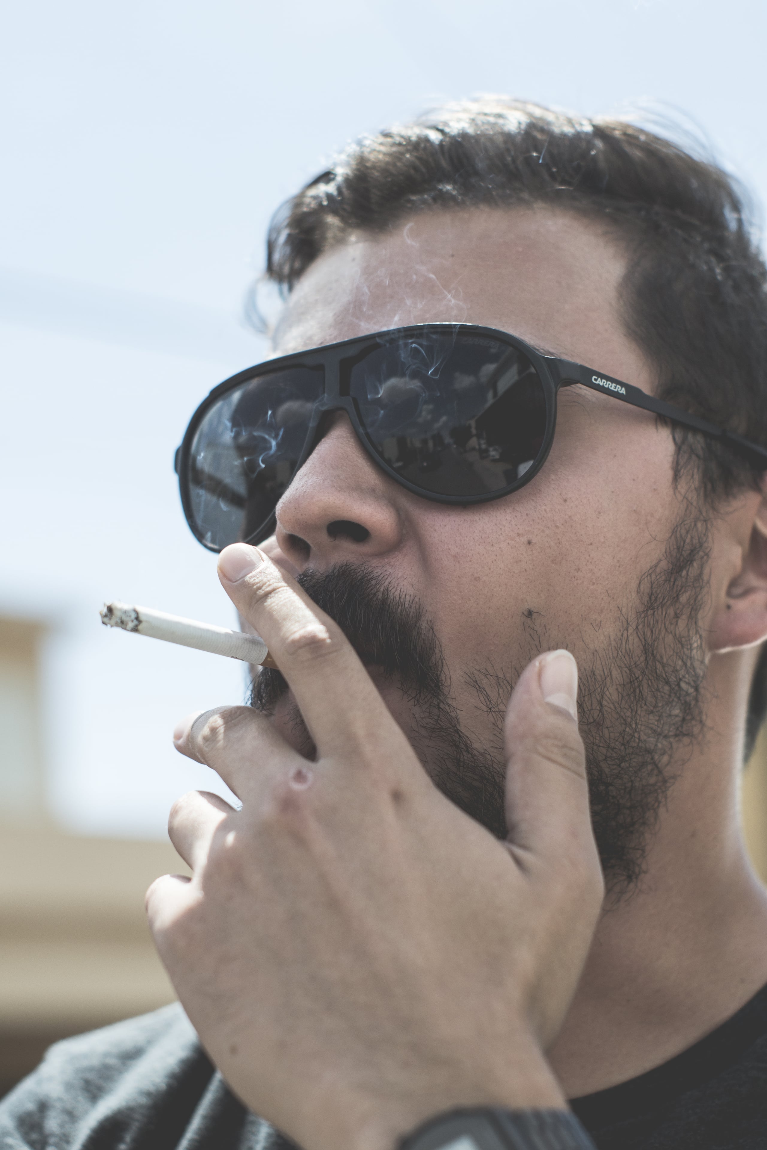 Close-up portrait of a bearded man wearing dark Carrera sunglasses smoking a cigarette, shot in a cinematic style with soft background blur