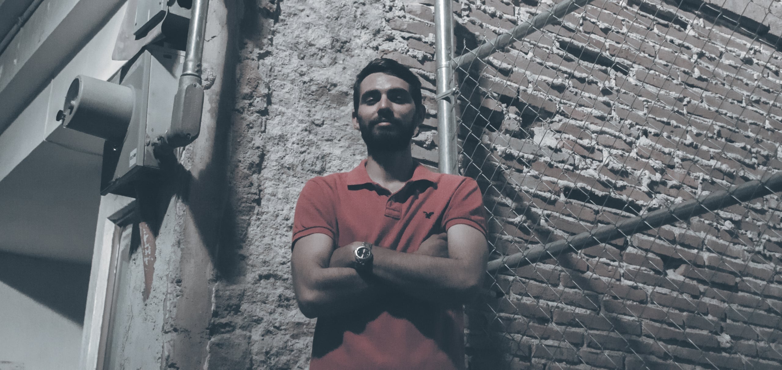 Portrait of a bearded man in a salmon-colored polo shirt standing with arms crossed in front of a textured brick wall and chain-link fence in an urban alley