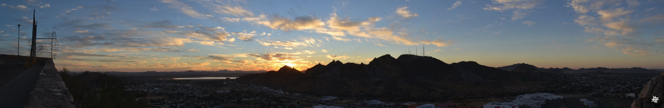 Panoramic sunset photograph showing mountain silhouettes with the sun setting behind peaks, golden and blue sky with scattered clouds over a city valley