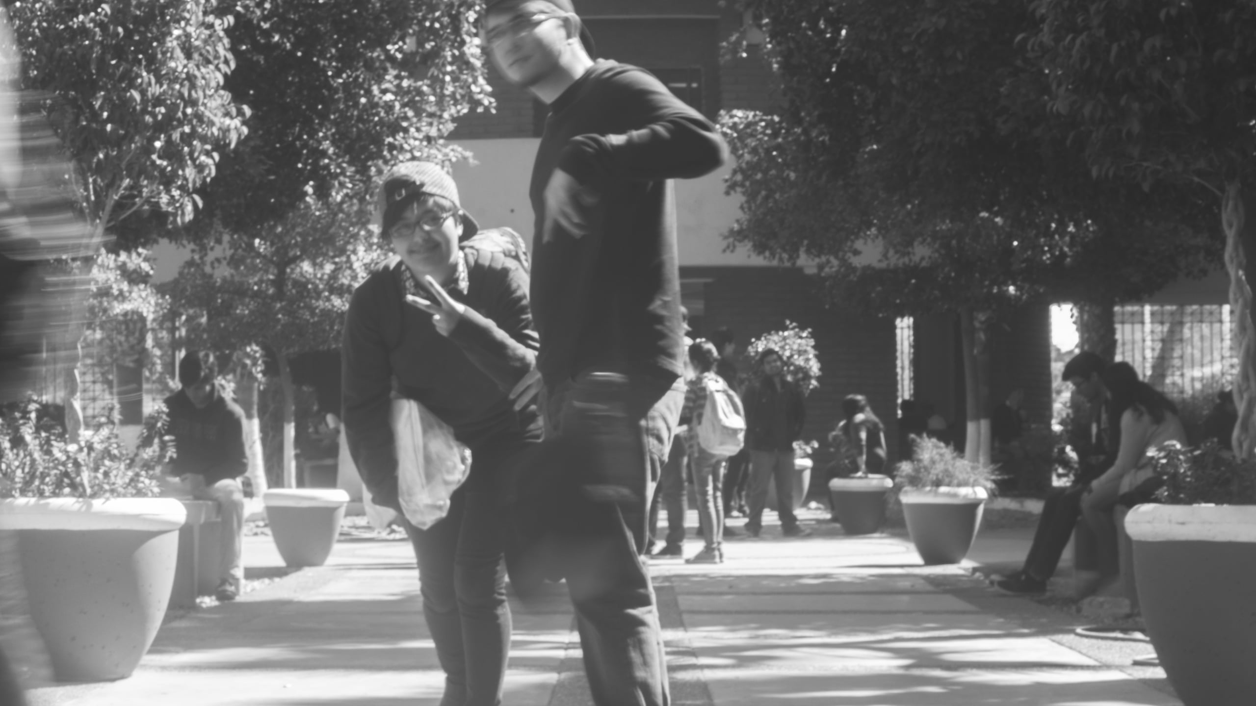 Black and white street photograph of two people standing in an outdoor plaza with trees, planters, and other pedestrians in the background