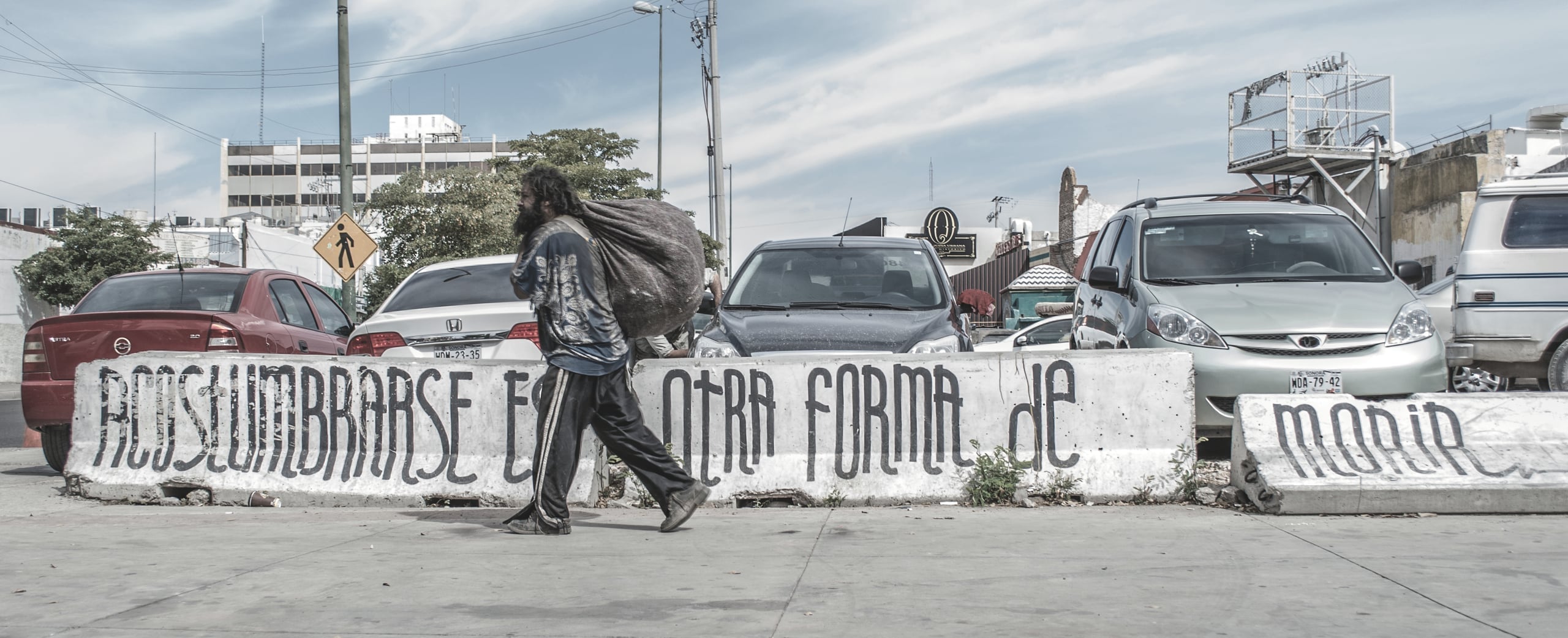 Street photograph of a person with long hair carrying a large bag walking past a concrete barrier with Spanish text graffiti, cars parked in background