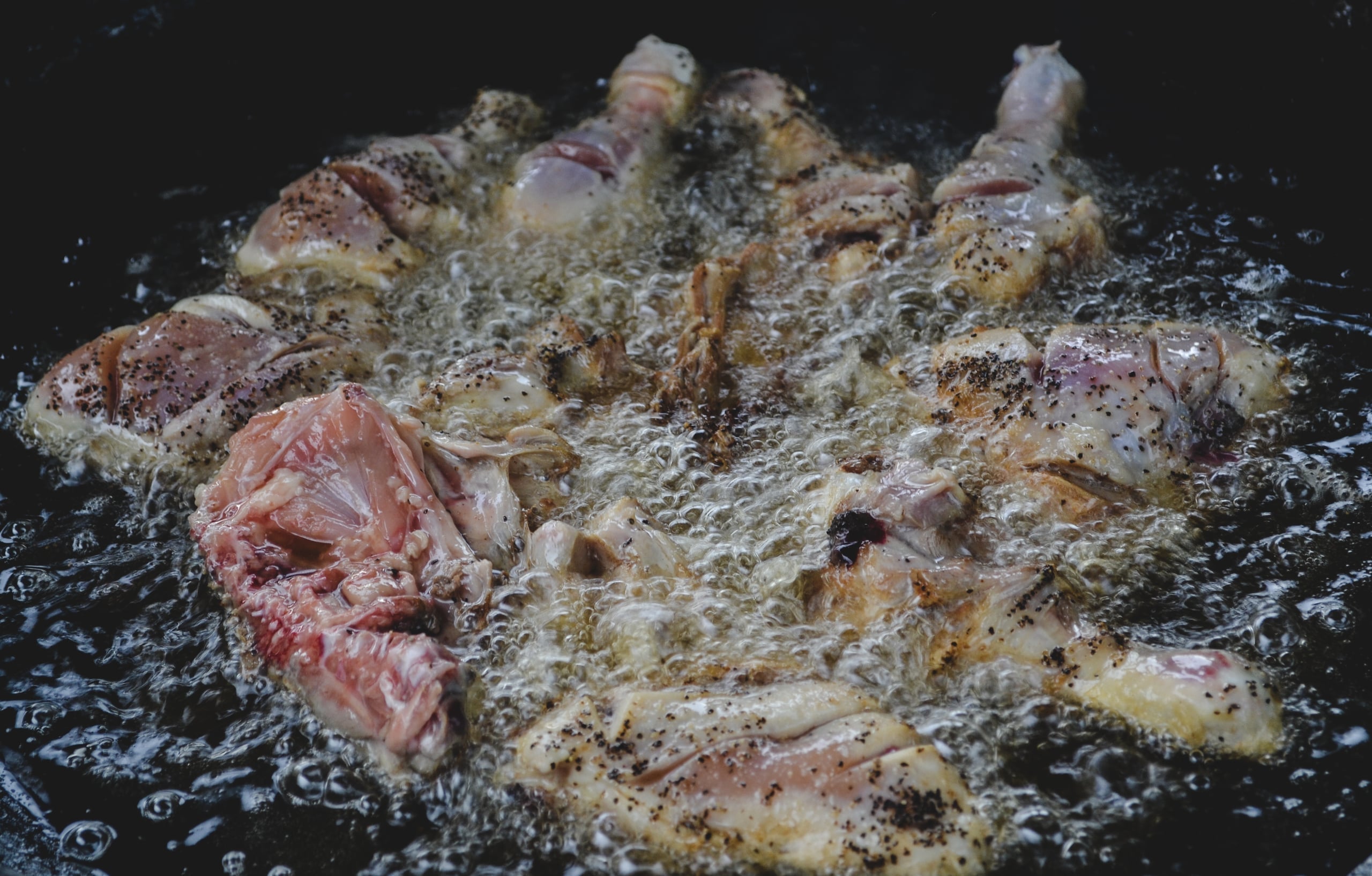 Close-up food photograph of seasoned chicken pieces frying in bubbling oil in a dark pan, showing texture and cooking process