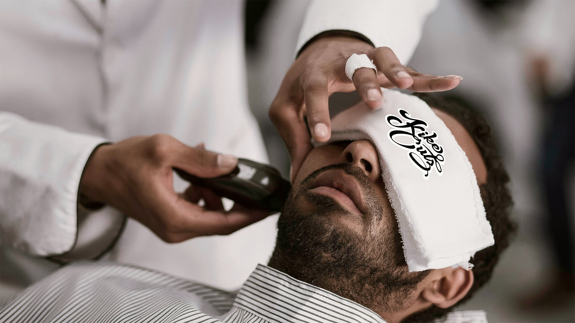 Close-up of barber trimming client beard with clippers while client reclines with branded Kike Cutz hot towel covering eyes in white salon environment