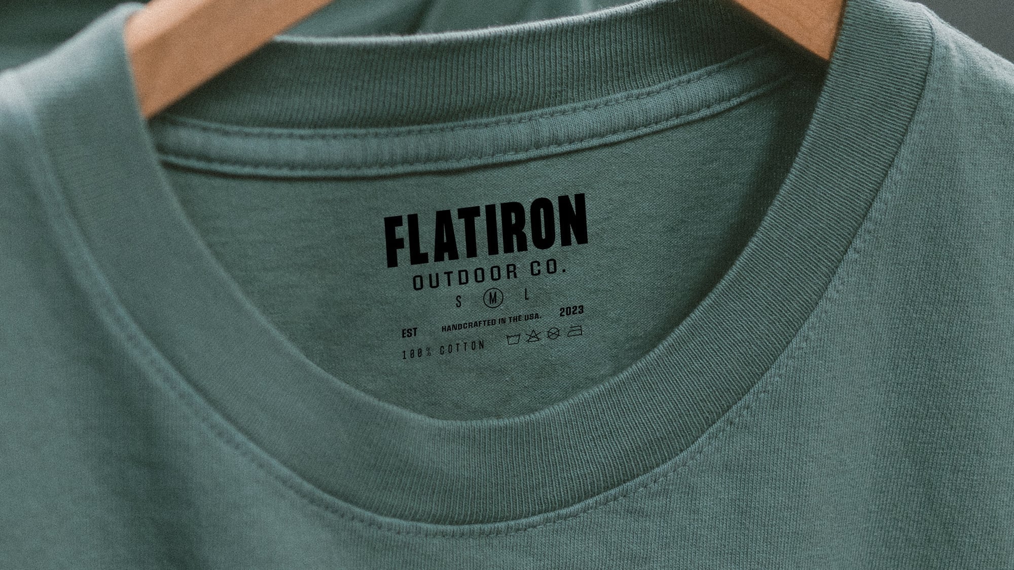 Close-up of sage green cotton t-shirt interior showing screen-printed neck label with Flatiron Outdoor Co. logo, size chart, and care symbols on wooden hanger.