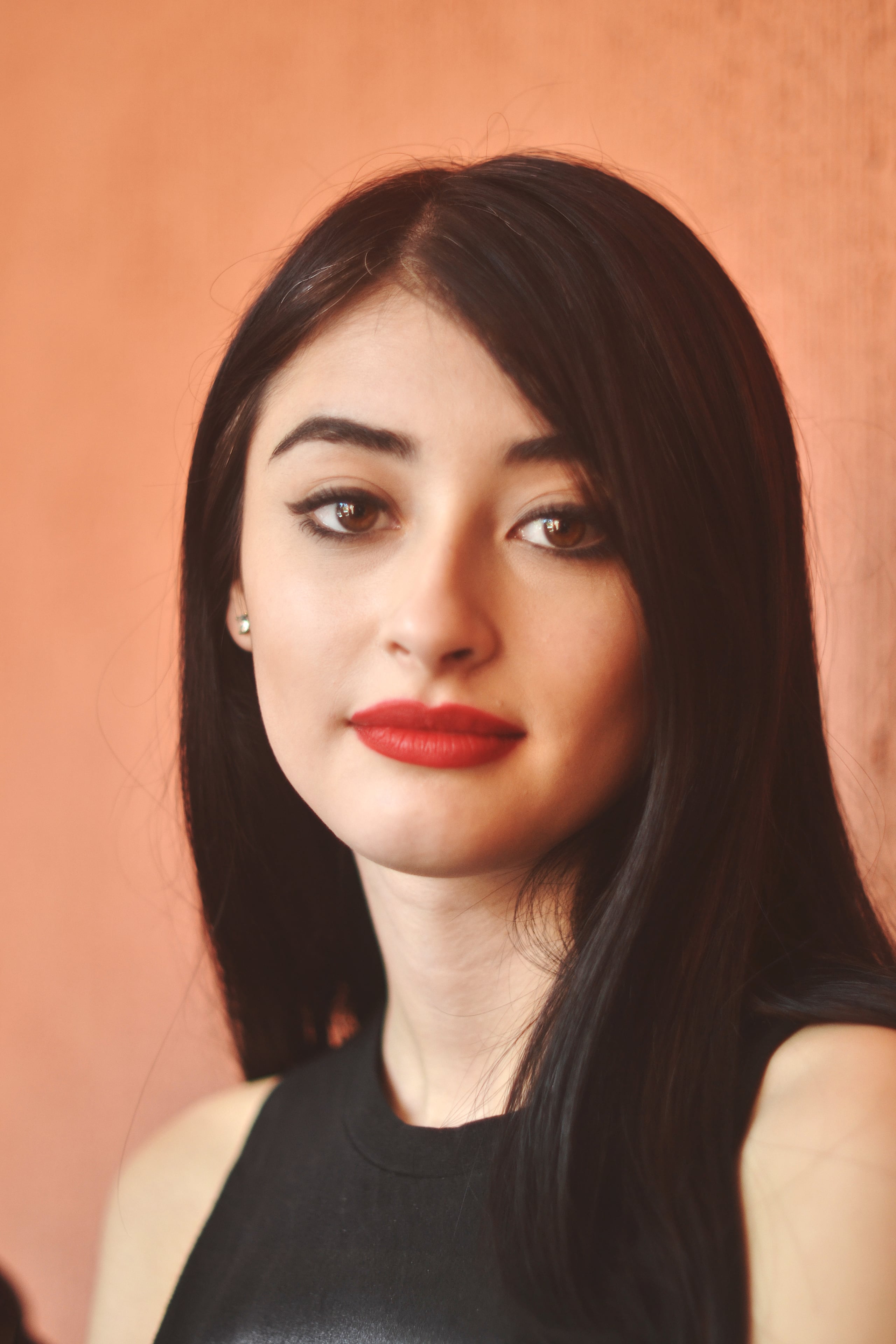 Studio portrait of a woman with long straight black hair and red lipstick against a warm peach-orange background, wearing a black sleeveless top