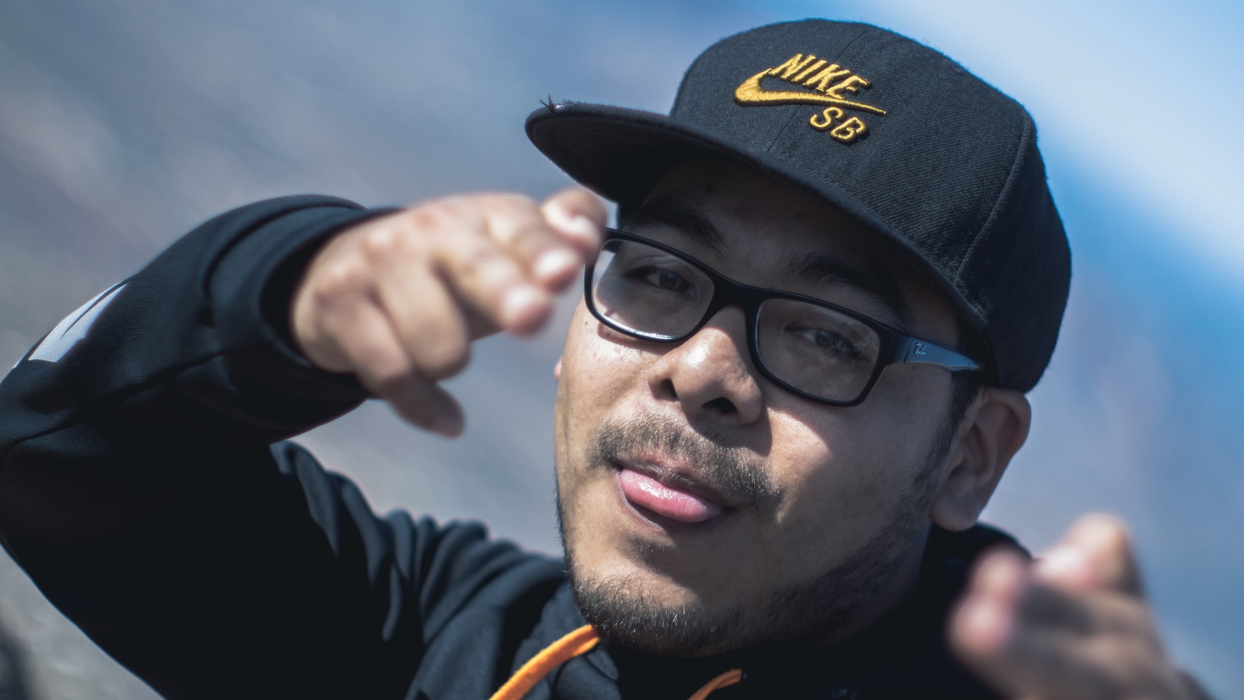 Close-up portrait of a man wearing a backwards black Nike cap and wire-rimmed glasses, with a slight mustache, shot against a bright sky