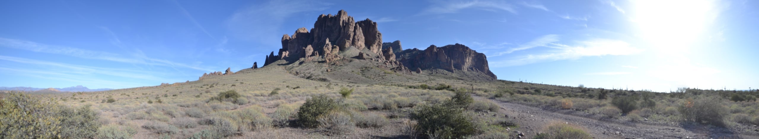 Panoramic landscape photograph of the Superstition Mountains in Arizona, jagged volcanic rock formations rising from desert scrubland under a blue sky