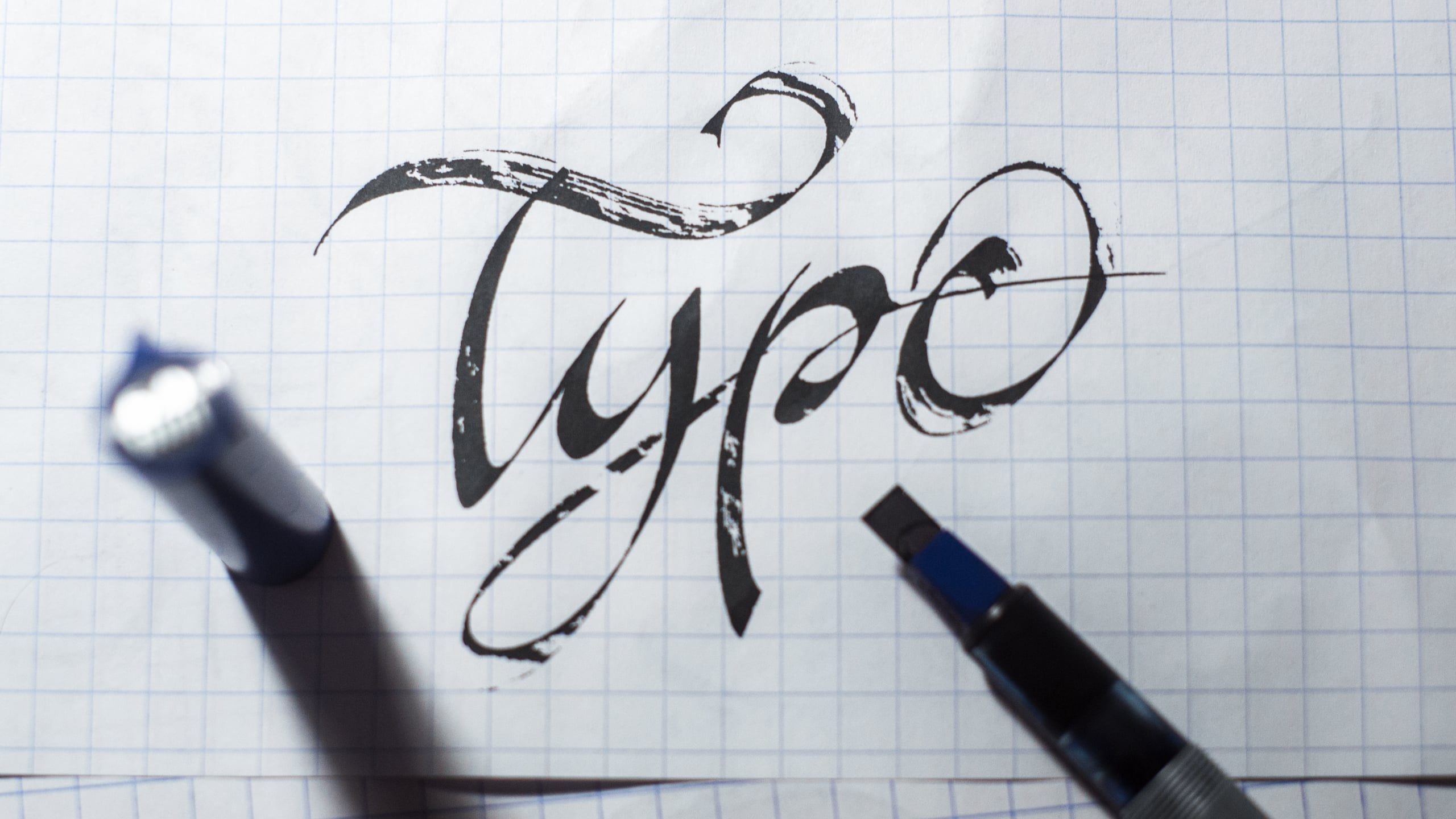 Hand-lettered calligraphy spelling 'Type' in bold black brush strokes on graph paper, with calligraphy pens visible at the edges
