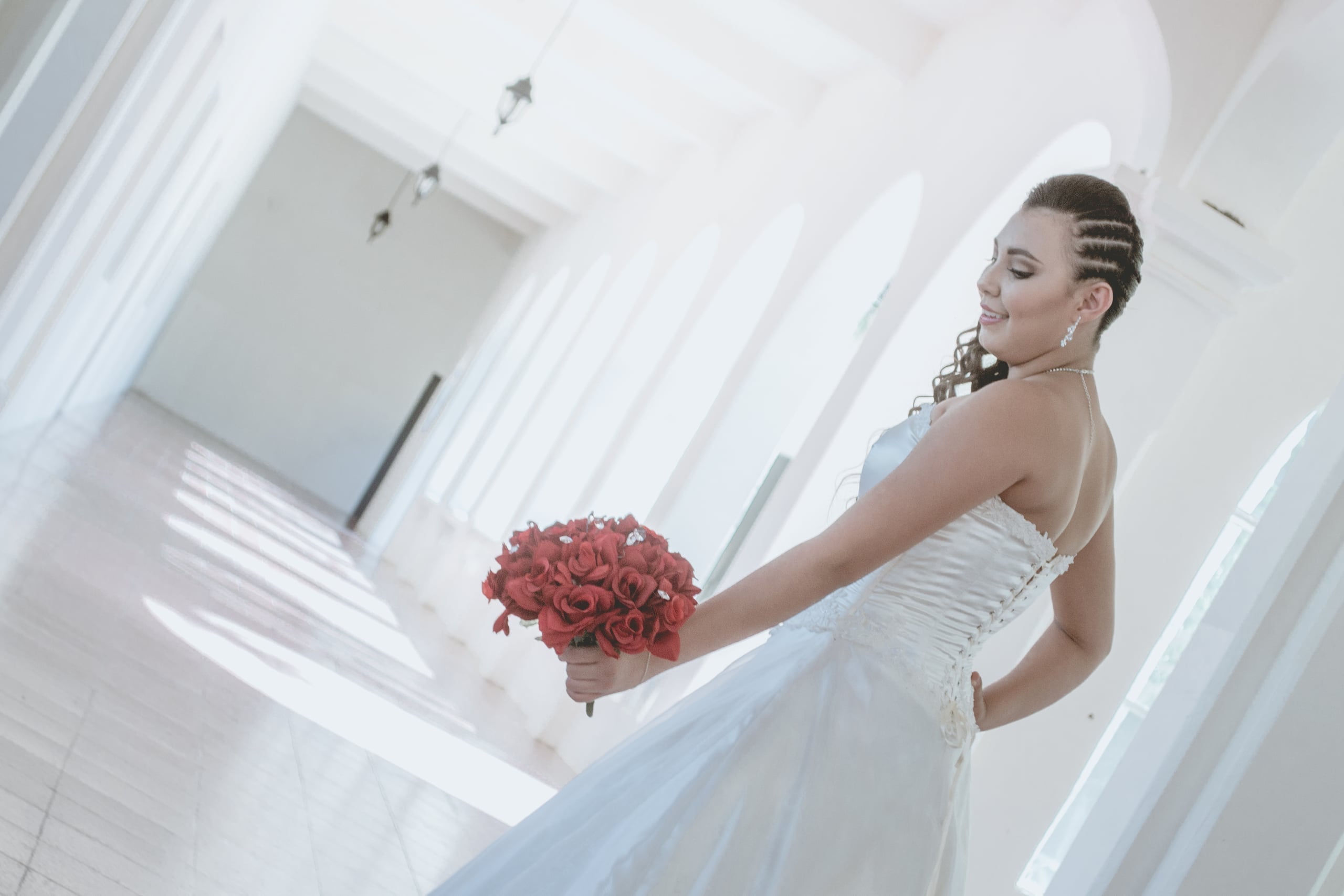 Elegant bridal portrait of a woman in a white wedding gown holding a bouquet of red roses, photographed in a bright white architectural space with arches