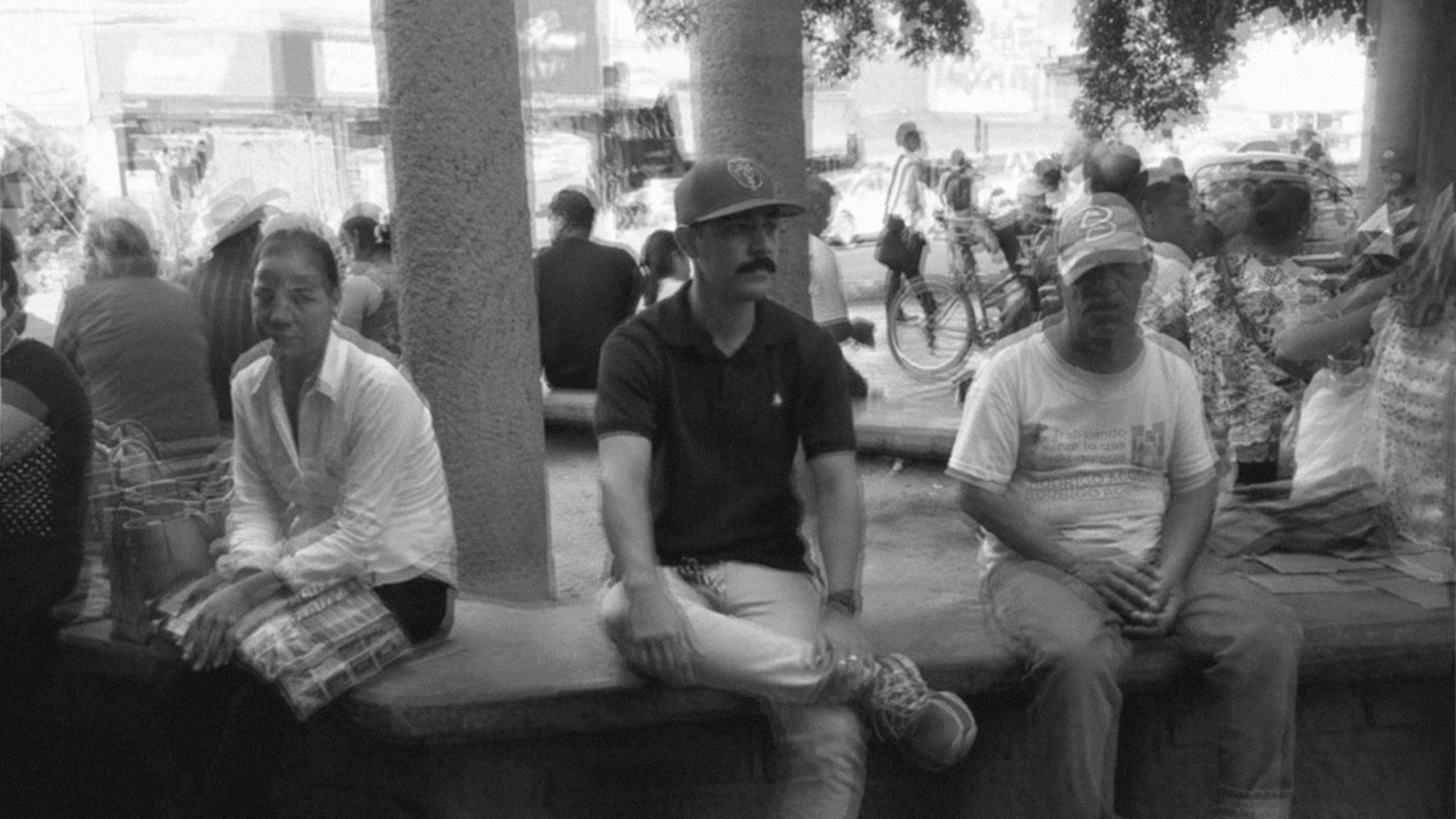 Black and white photo of Raúl René Romero Ramos, graphic designer, sitting on a bench in a Mexican town plaza wearing a polo shirt and cap, with locals nearby.