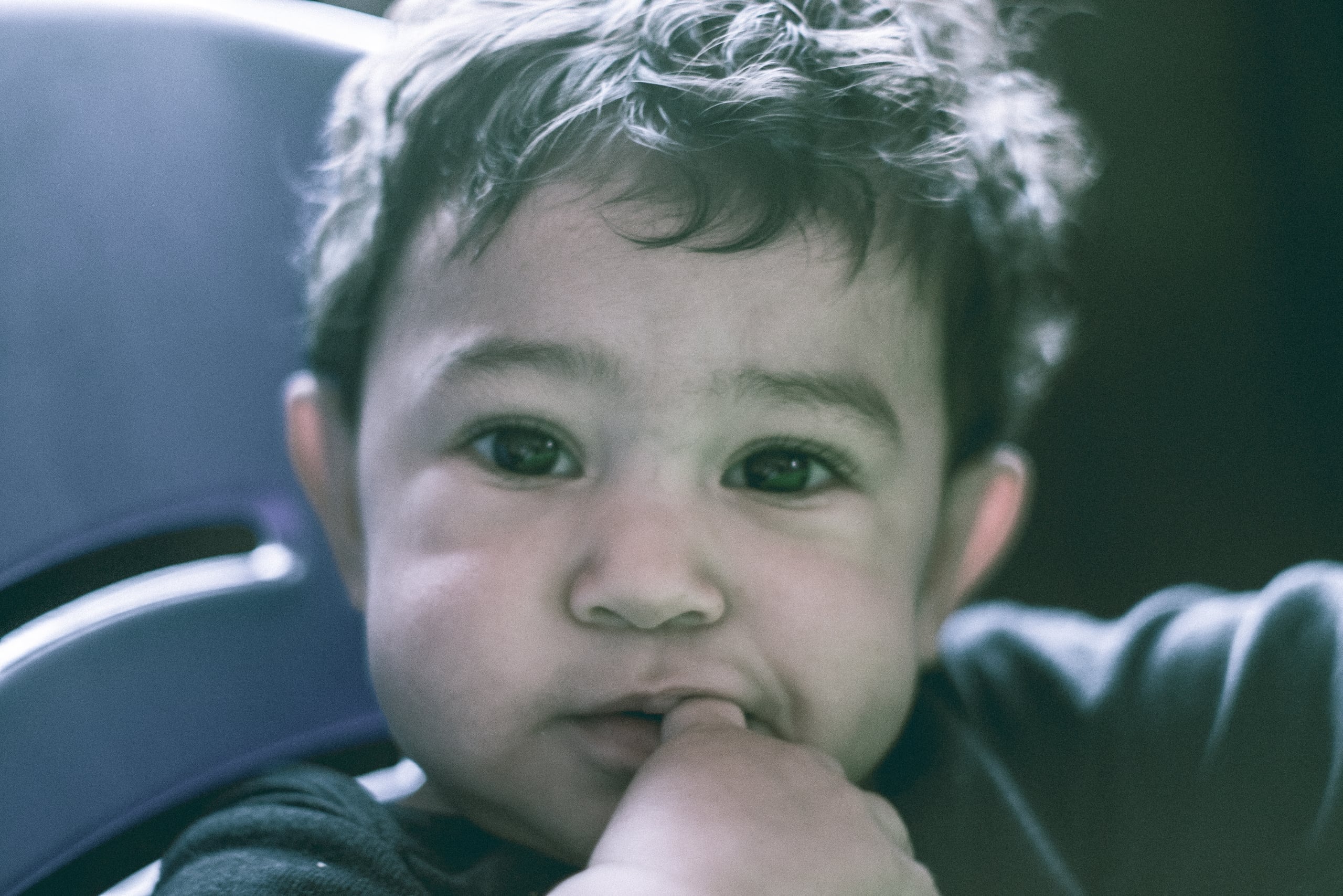Intimate close-up portrait of a curly-haired toddler with big brown eyes sucking their thumb, sitting in a car seat with cinematic color grading