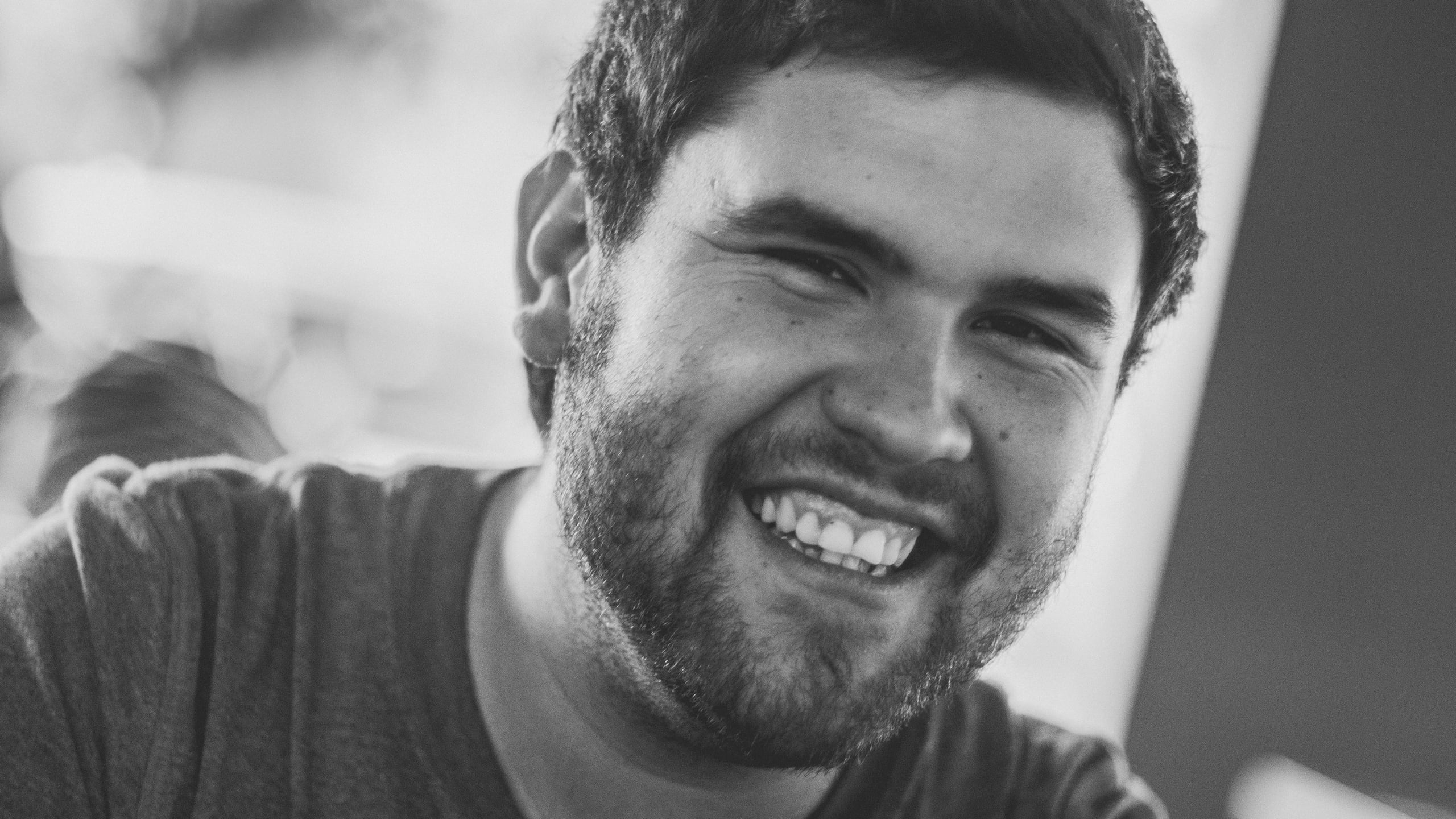 Black and white close-up portrait of a young bearded man laughing genuinely, wearing a casual t-shirt with soft natural lighting