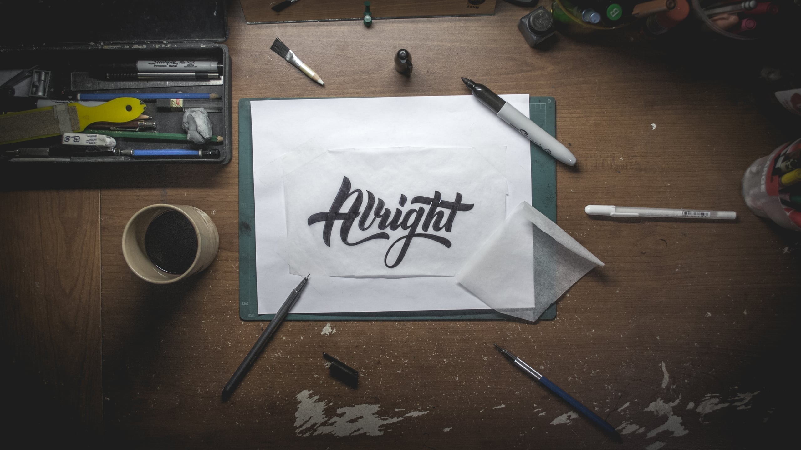 Overhead view of a creative workspace with black brush lettering spelling 'Alright' on paper, surrounded by pens, markers, coffee cup, and art supplies