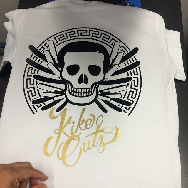 White t-shirt with black skull and barber comb design surrounded by Greek key pattern with gold Kike Cutz script below