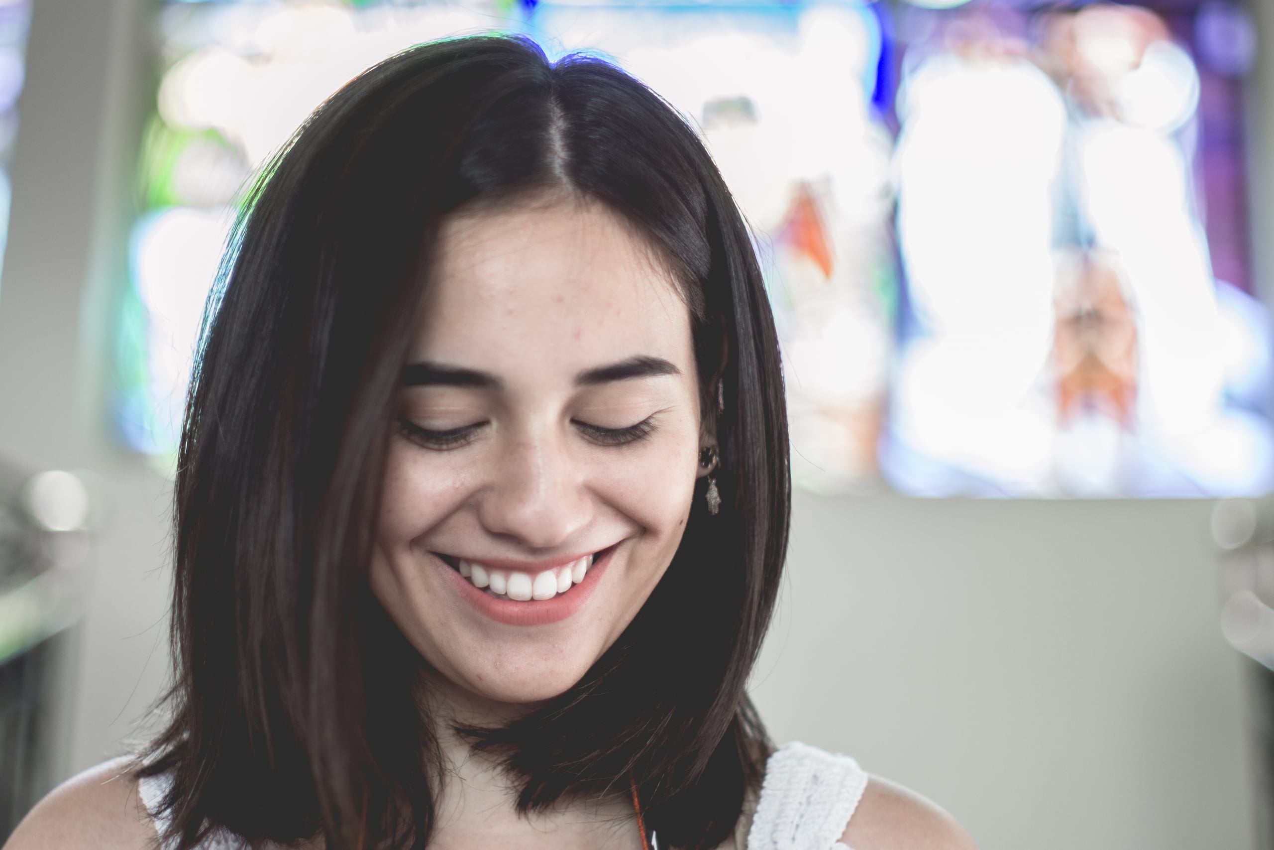 Portrait of a young woman with dark shoulder-length hair and eyes closed, smiling peacefully, with colorful stained glass window bokeh in the background