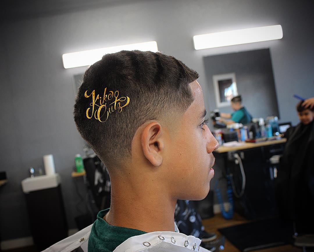 Side profile of young male client showcasing fresh fade haircut with gold Kike Cutz watermark logo overlay in barbershop setting