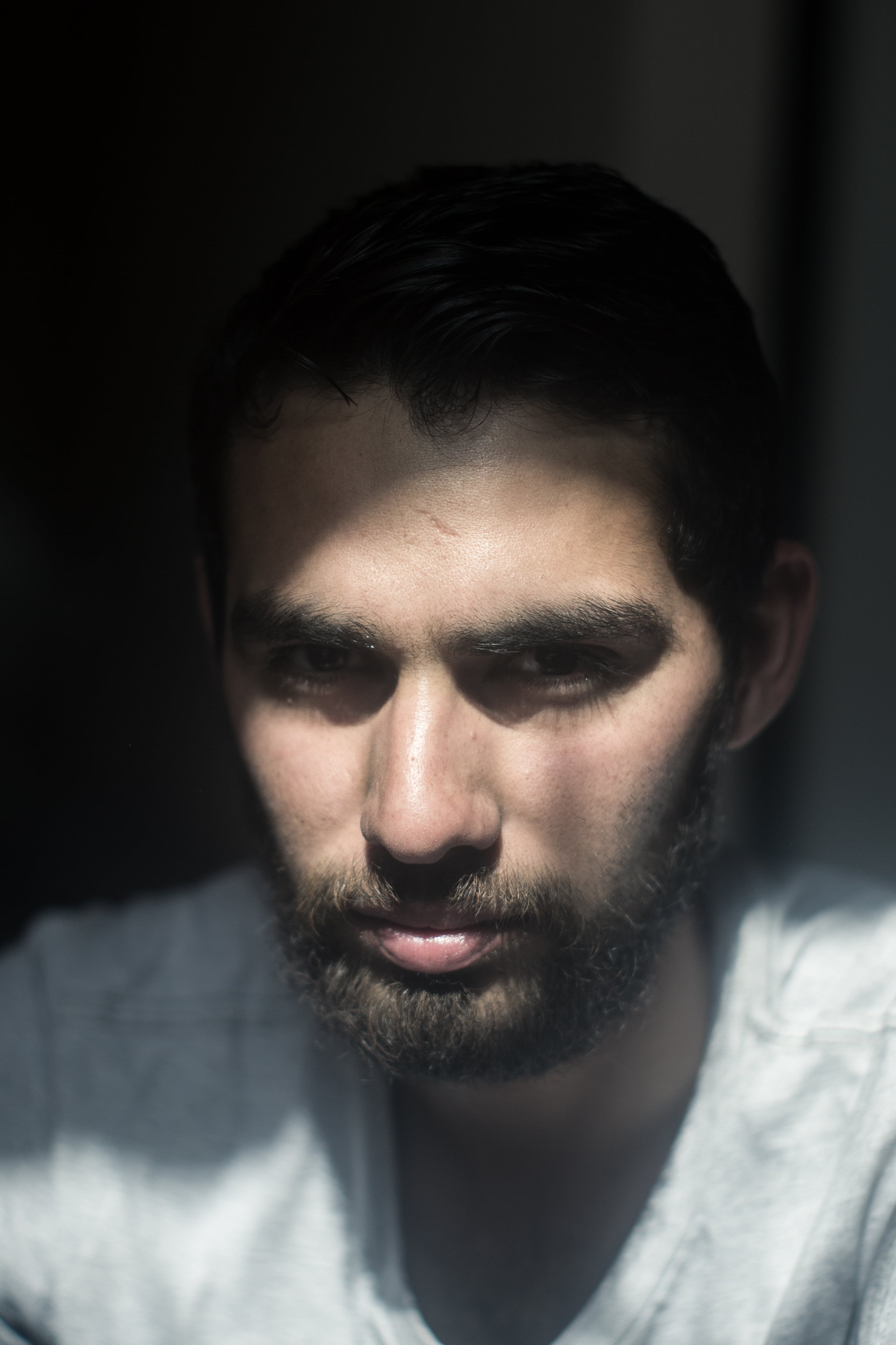 Dramatic portrait of a bearded man with strong shadows across his face from window light, creating a chiaroscuro effect in muted tones