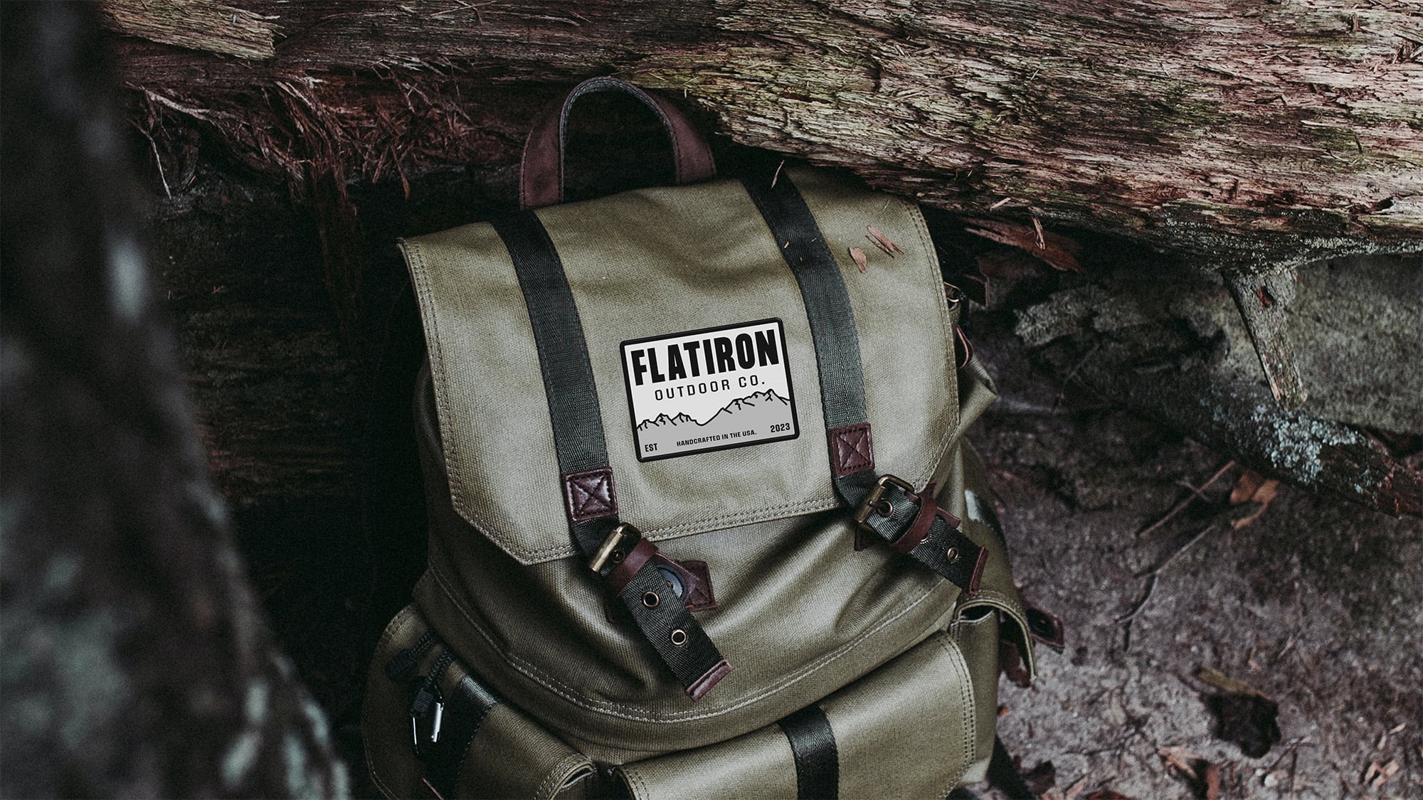 Vintage-style khaki canvas rucksack with dark leather straps and brass buckles, featuring Flatiron Outdoor Co. mountain logo patch, photographed against weathered tree bark in forest setting.