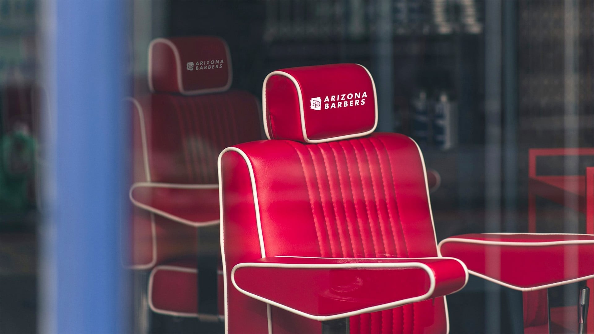 Red leather barber chairs with white piping featuring embroidered Arizona Barbers logo on headrests, viewed through barbershop window