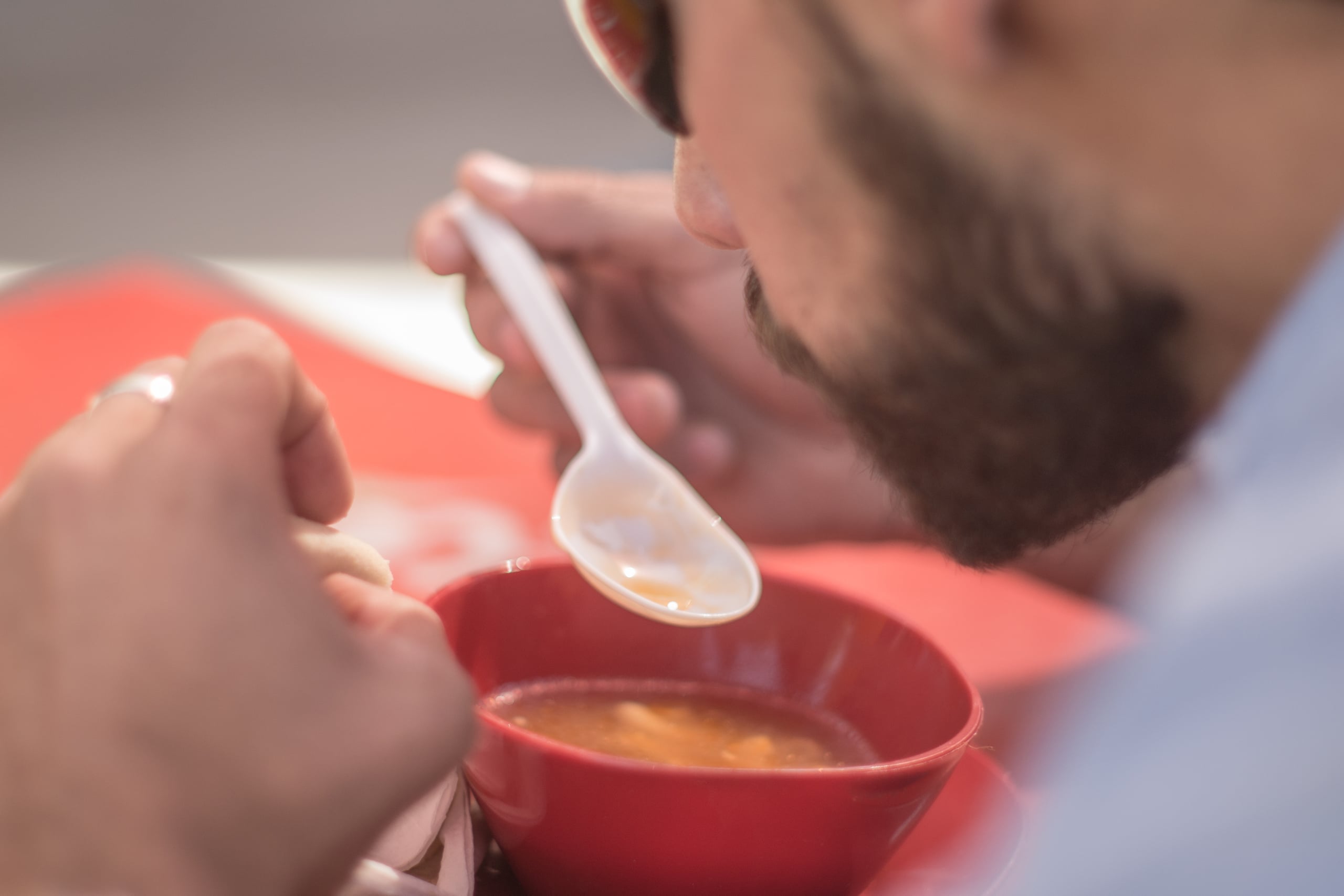 Close-up of a bearded man eating soup from a red ceramic bowl with a white plastic spoon, shot from the side with shallow depth of field
