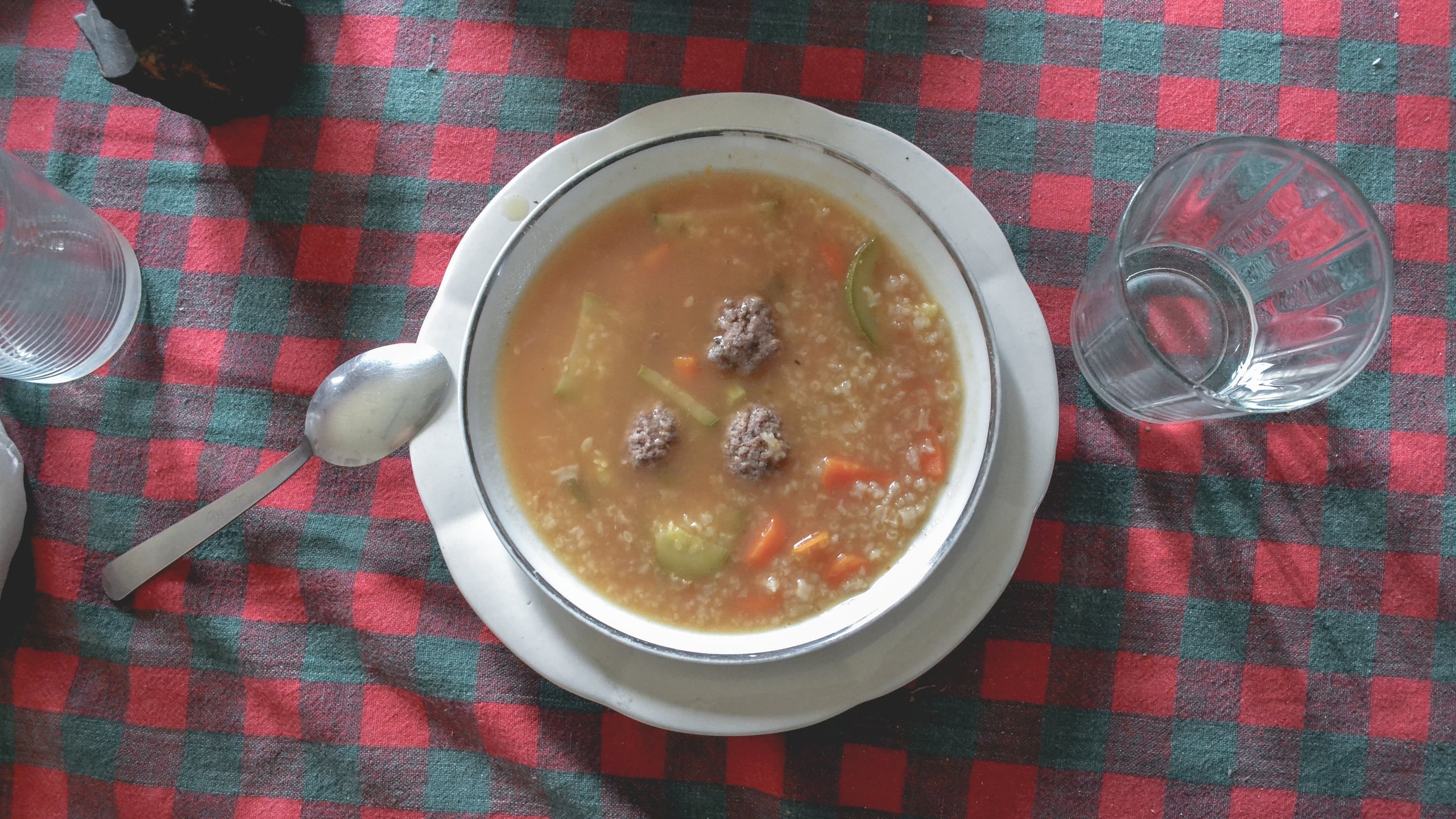 Overhead food photograph of a hearty vegetable soup with meatballs in a white bowl on a red and green checkered tablecloth, with spoon and water glass