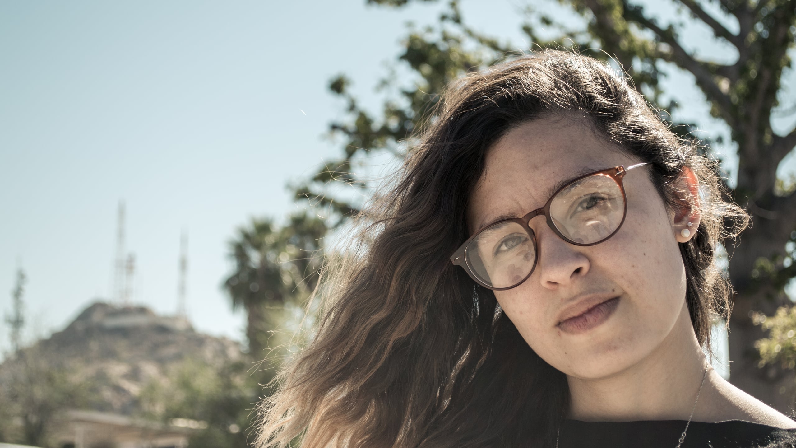 Close-up portrait of a young woman with long wavy brown hair and round glasses, looking directly at the camera outdoors with trees and hillside behind her