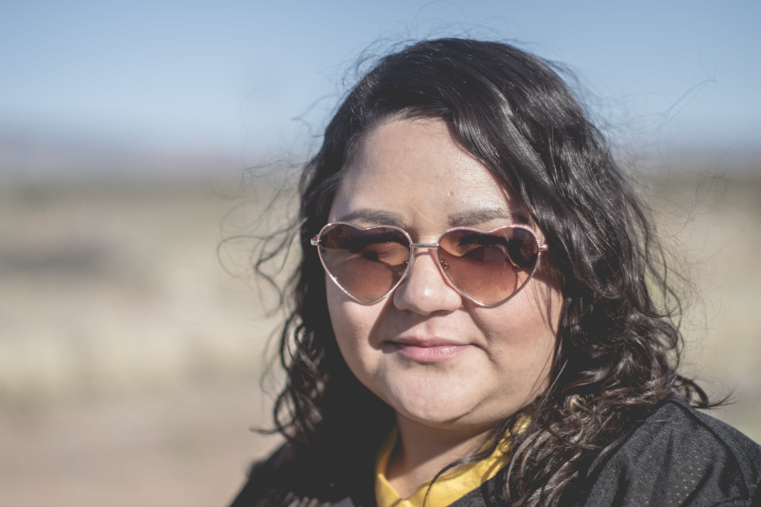Portrait of a woman with dark wavy hair wearing heart-shaped pink sunglasses, shot outdoors against a blurred desert landscape