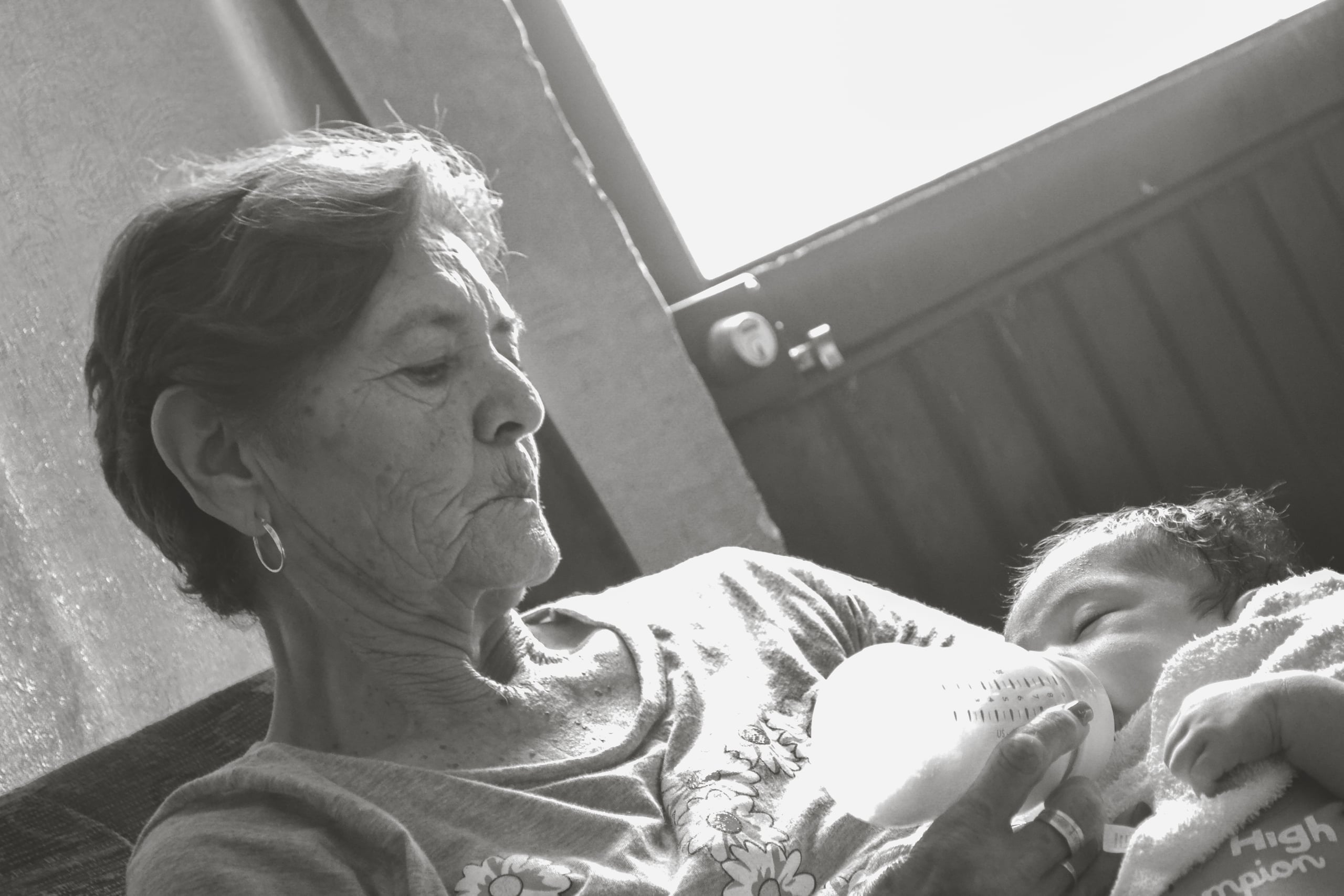 Tender black and white photograph of an elderly grandmother bottle-feeding an infant, showing weathered hands and intimate family connection