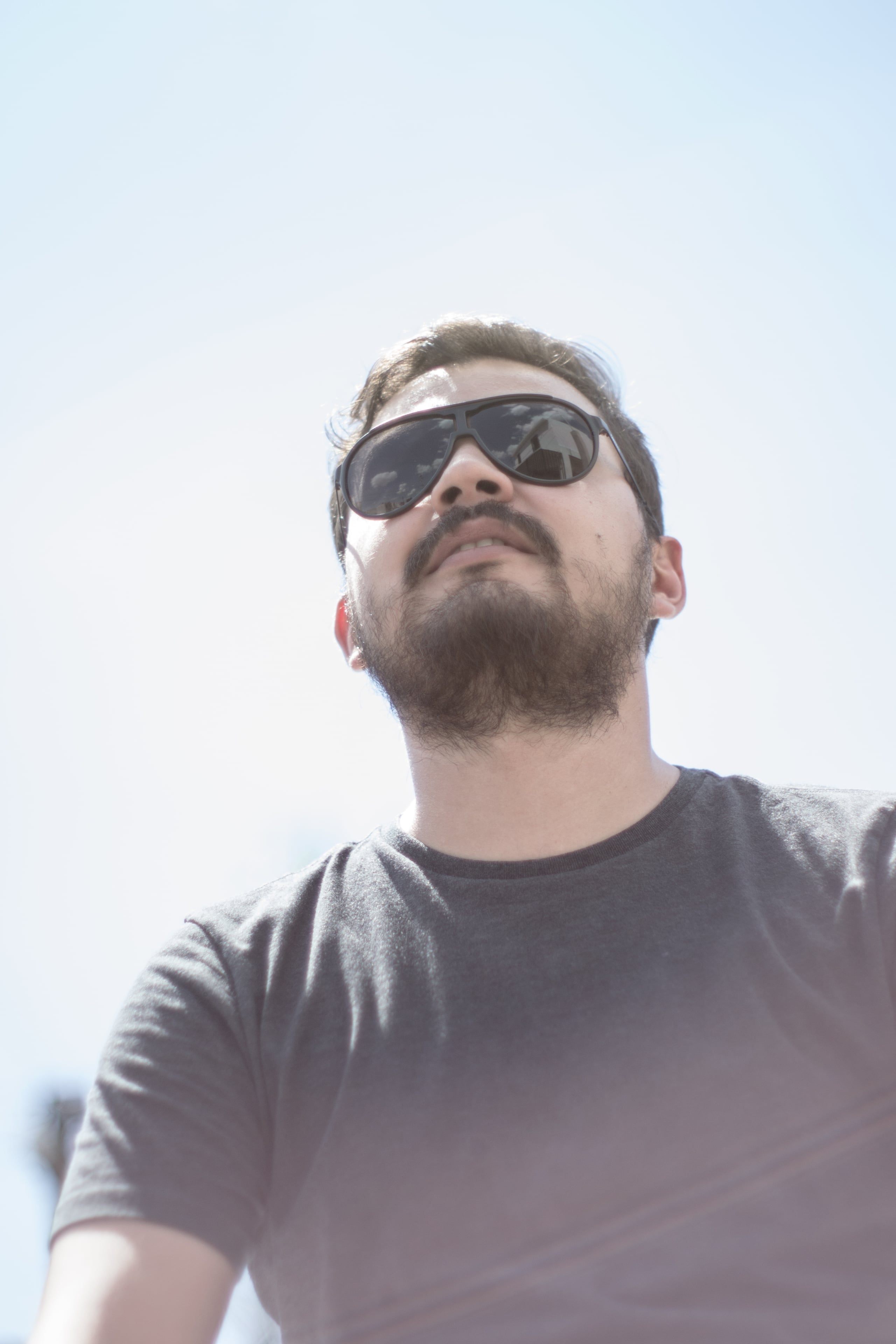 Low angle portrait of a bearded man wearing aviator sunglasses and a gray t-shirt, looking upward against a bright overexposed sky